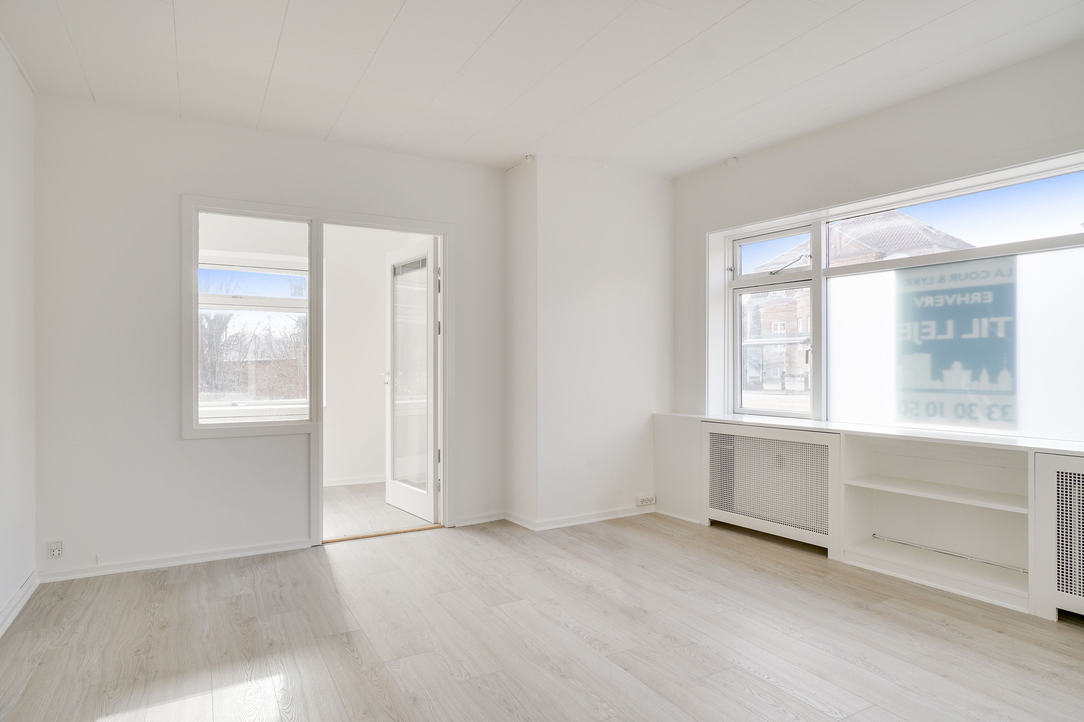 Kontorrum med hvide vægge og gulv samt glasdøre og vinduer, radiatorer og højhyggeligt lys fra vinduet, og et lejlighedsvist glasskilt med engelsk tekst