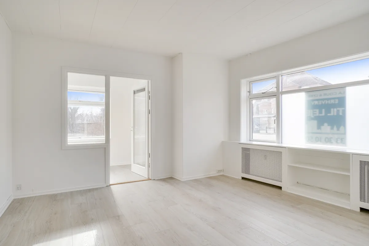 Kontorrum med hvide vægge og gulv samt glasdøre og vinduer, radiatorer og højhyggeligt lys fra vinduet, og et lejlighedsvist glasskilt med engelsk tekst