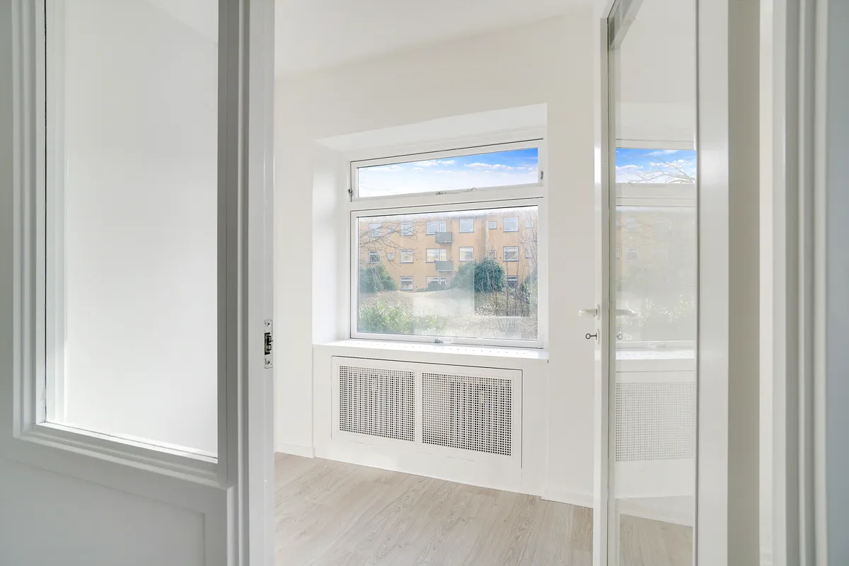 Lukket hvidt rum med vinduesåbninger og hvidt gulv, der vender mod en bygning med solrig himmel og træer i baggrunden