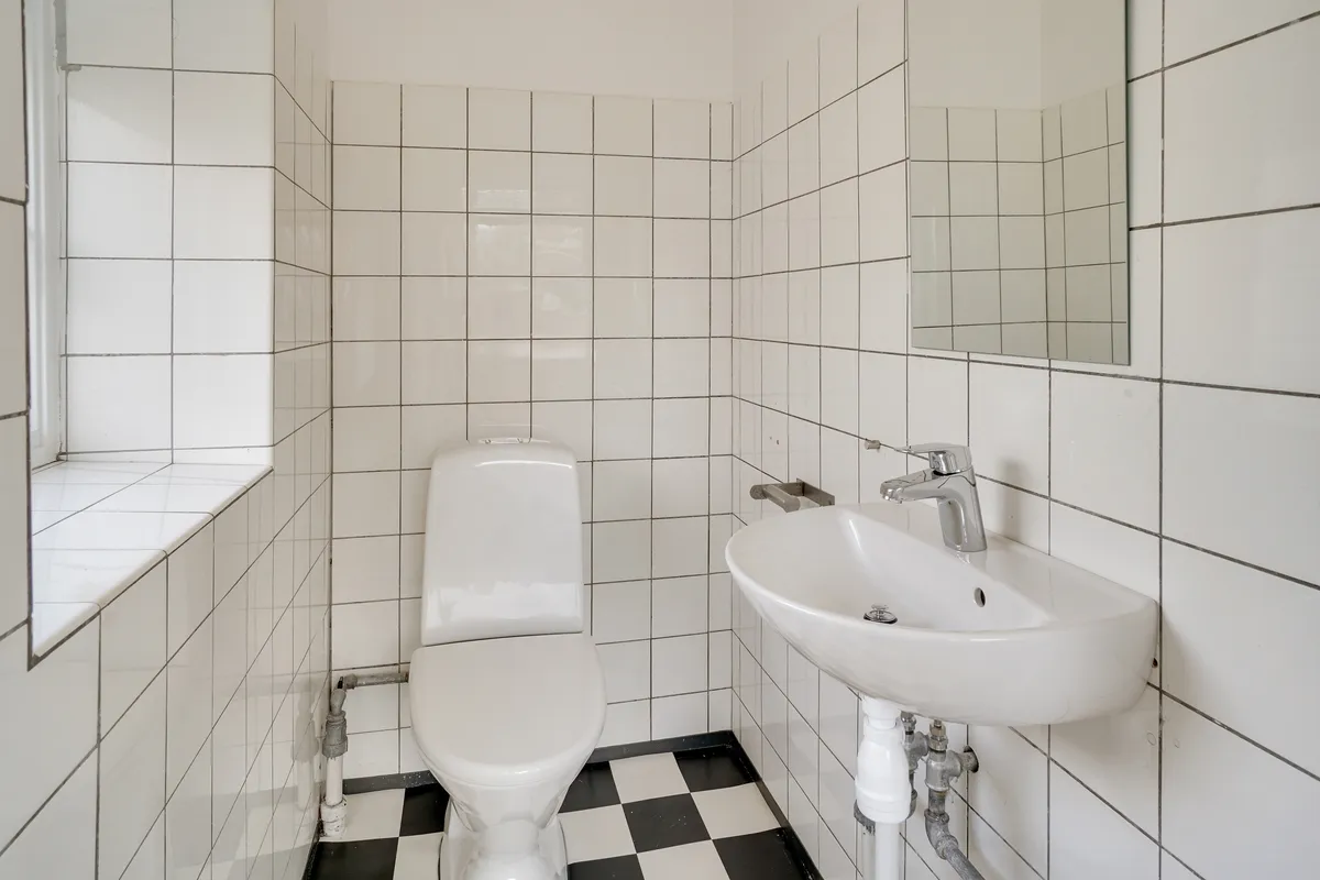 En lille hvid toilet i en hvidt tøjet toiletrum med hvidt spejl og hvidt vask. Sort og hvid gulvbelægning.