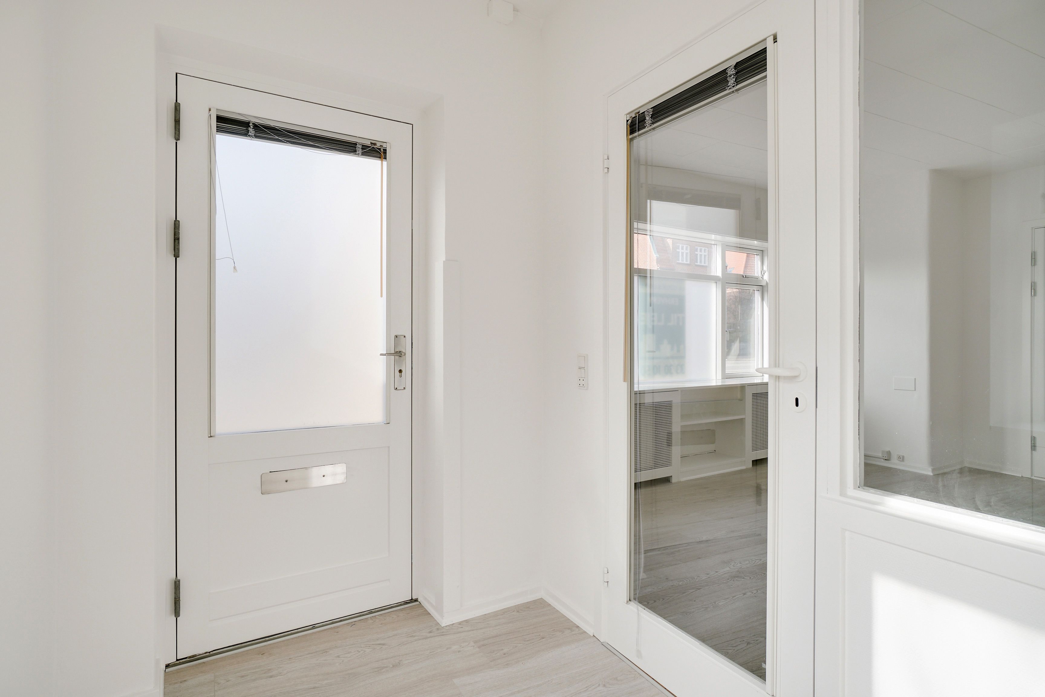 Kontor med hvide vægge og hvidt gulv, hvor døre og vinduer er åbne, og der er et spejl i det ene værelse, der reflekterer rummet