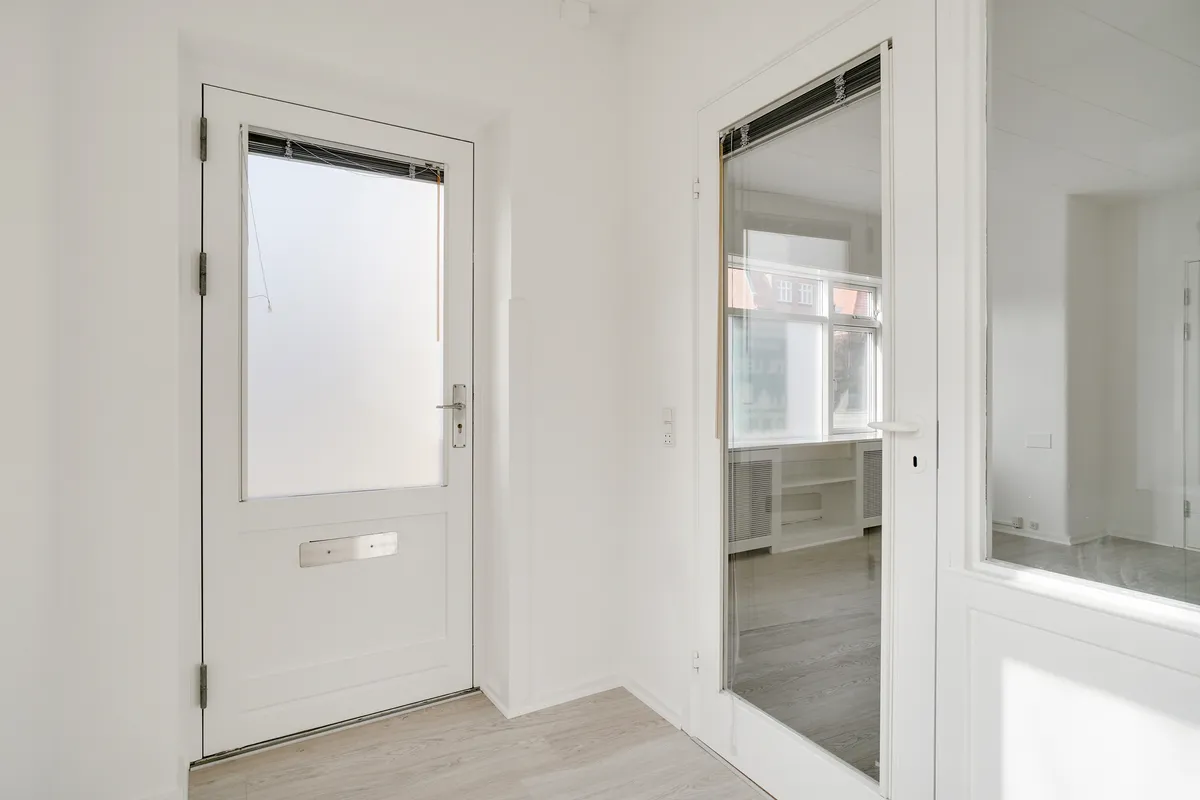 Kontor med hvide vægge og hvidt gulv, hvor døre og vinduer er åbne, og der er et spejl i det ene værelse, der reflekterer rummet