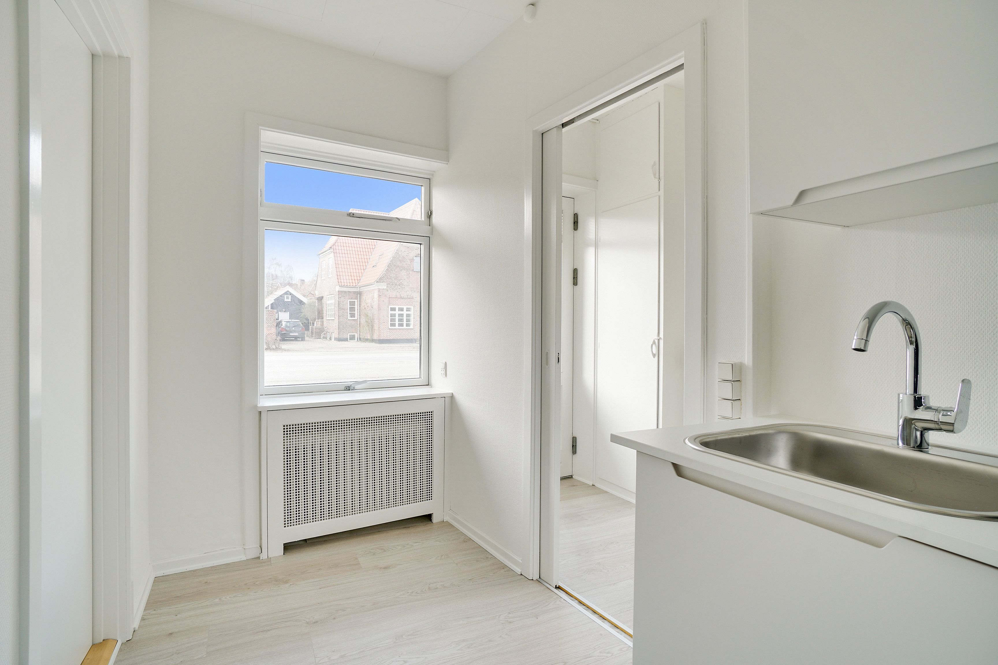 Rent kontor med hvid væg, hårdt trægulv og lys. Simpel køkkenudstyret med en høj, enkel vask og vandhane. Vindue med udsigt til gade og bygninger.