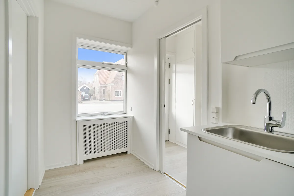 Rent kontor med hvid væg, hårdt trægulv og lys. Simpel køkkenudstyret med en høj, enkel vask og vandhane. Vindue med udsigt til gade og bygninger.