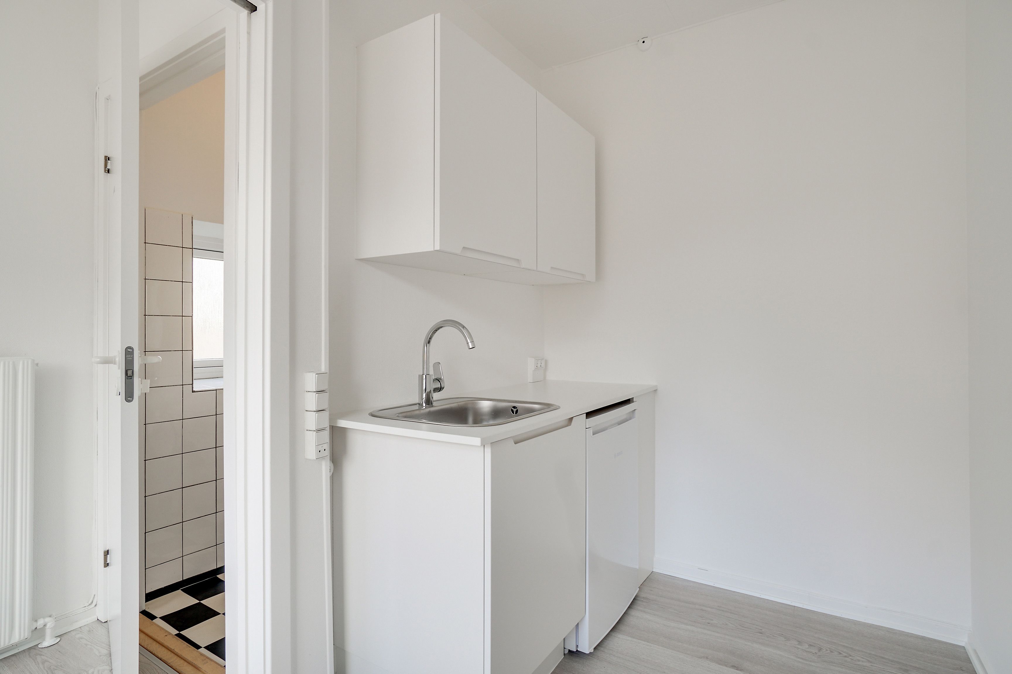 Kontor med hvidt køkken og sort/hvidt gulvtæppe. Rummet har hvidt loft og vægge, køkken med lysegråt loft og hvid keramik. Værelset har en radiator og hvidt gulv