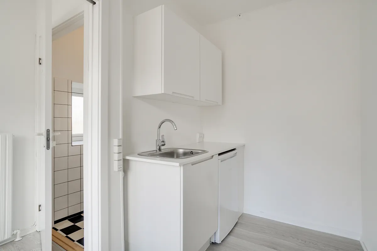 Kontor med hvidt køkken og sort/hvidt gulvtæppe. Rummet har hvidt loft og vægge, køkken med lysegråt loft og hvid keramik. Værelset har en radiator og hvidt gulv