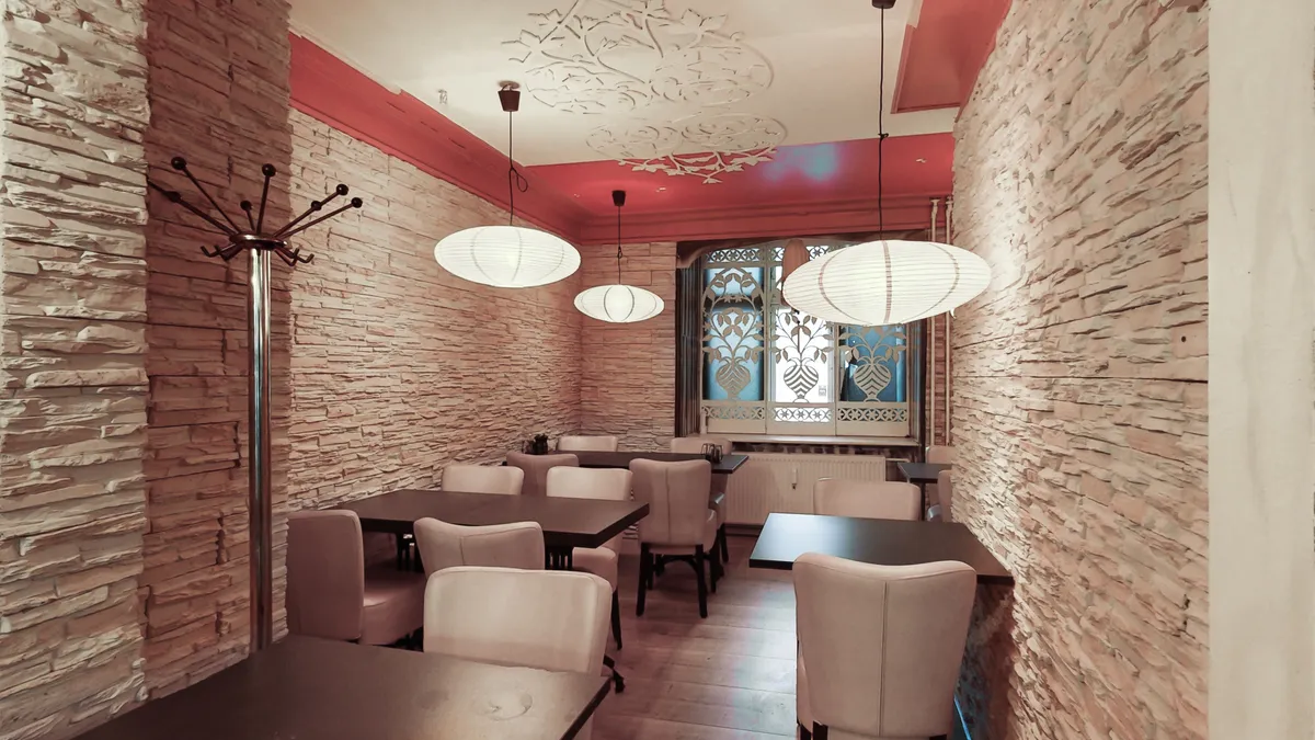 Inde i restaurant med stenvægge, hvide stolhede og runde paplamper fra loftet. Bord og stole er stillet, og der er et vindue med mønstret glas.