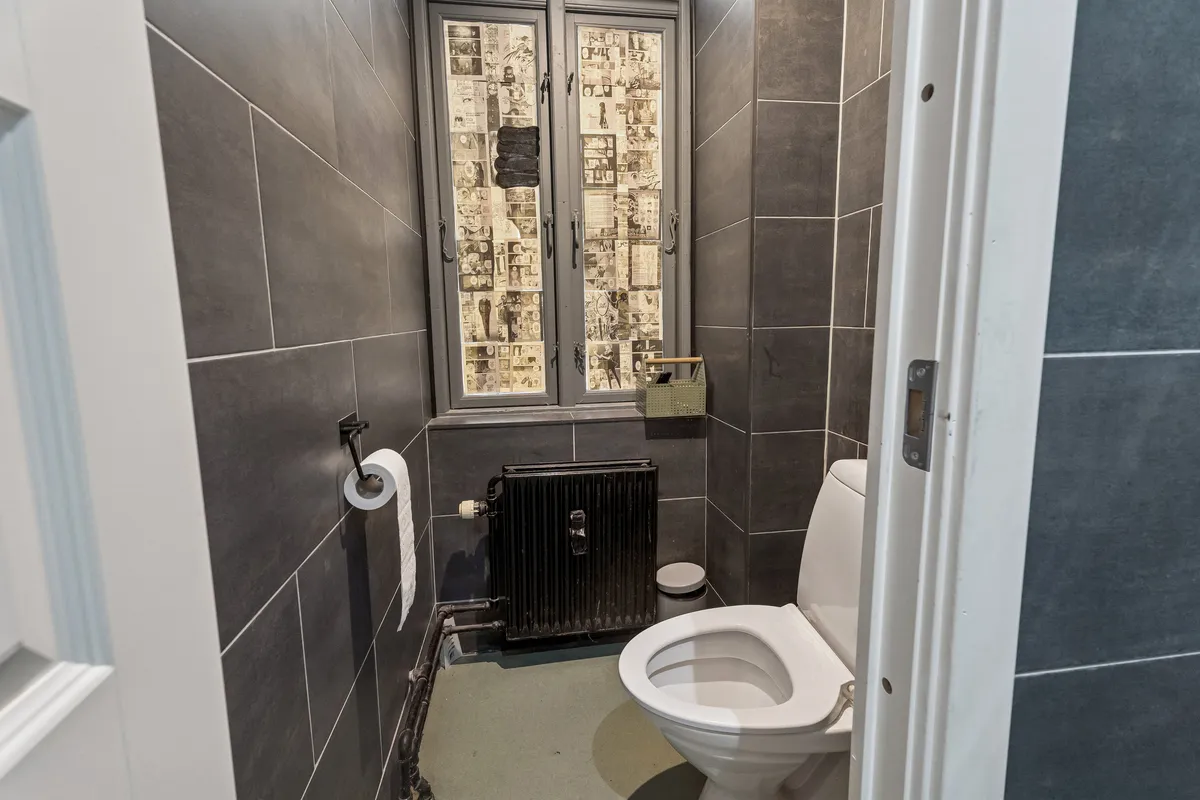 En moderne og veludstyret toiletrum med hvide vægge, en hvid toilet med åben dør, toiletpapir og en radiator i hjørnet, og en spejltørrer med to tørklæder