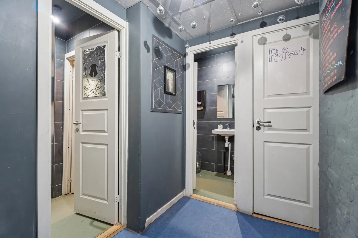 To rum med hvide døre og blå gulvbelægning, et badeværelse med spejl, håndklæder og en hvid vask, et vægmaleri med et billede og et sort tavle med tekst