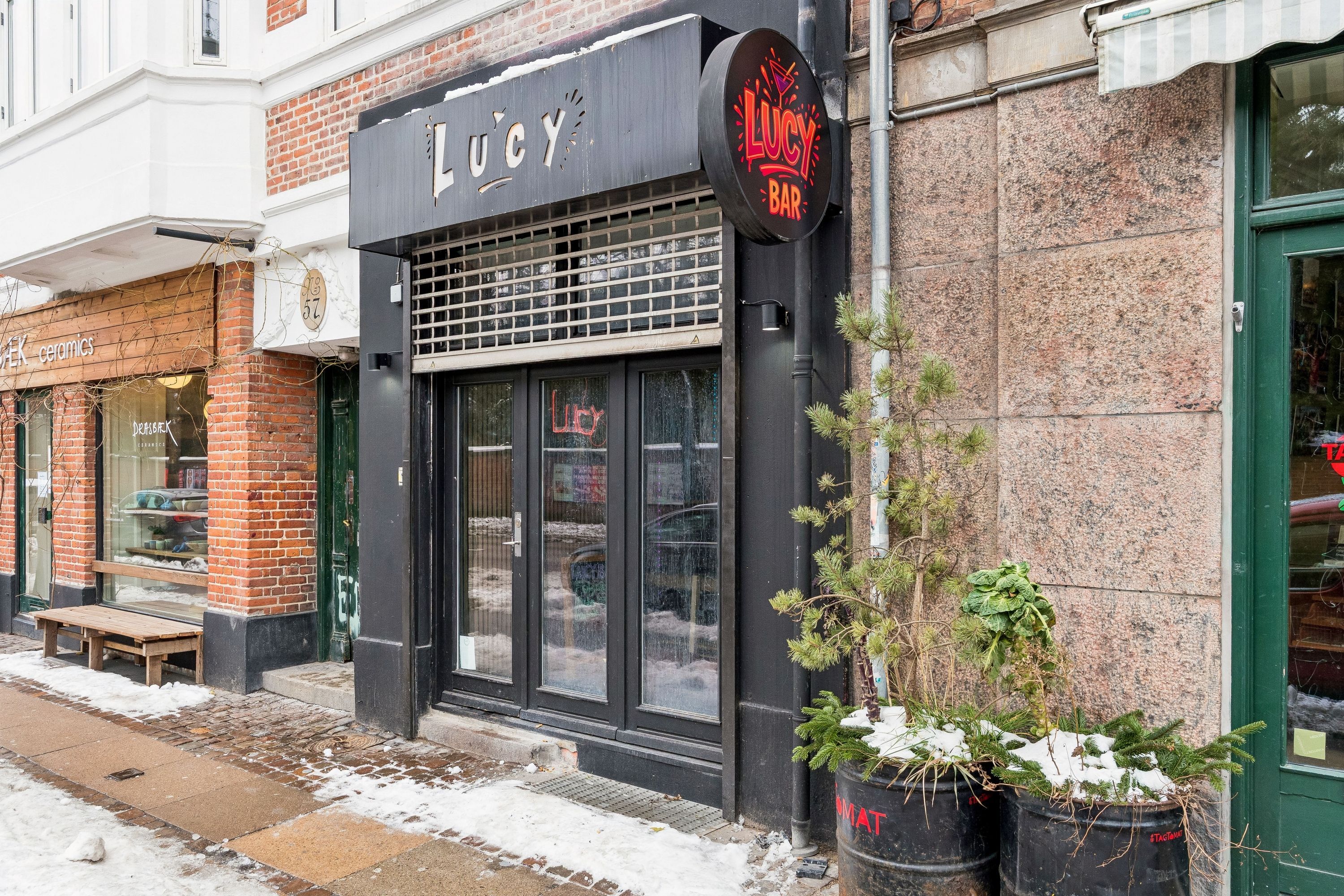Bar Lucy til leje på Jagtvej 57 i København. Bar med glasdøre og en udsmykning af sne-dækkede planter udenfor. Beliggenhed i et koldt vinterlandskab med sne på gaden og bygninger