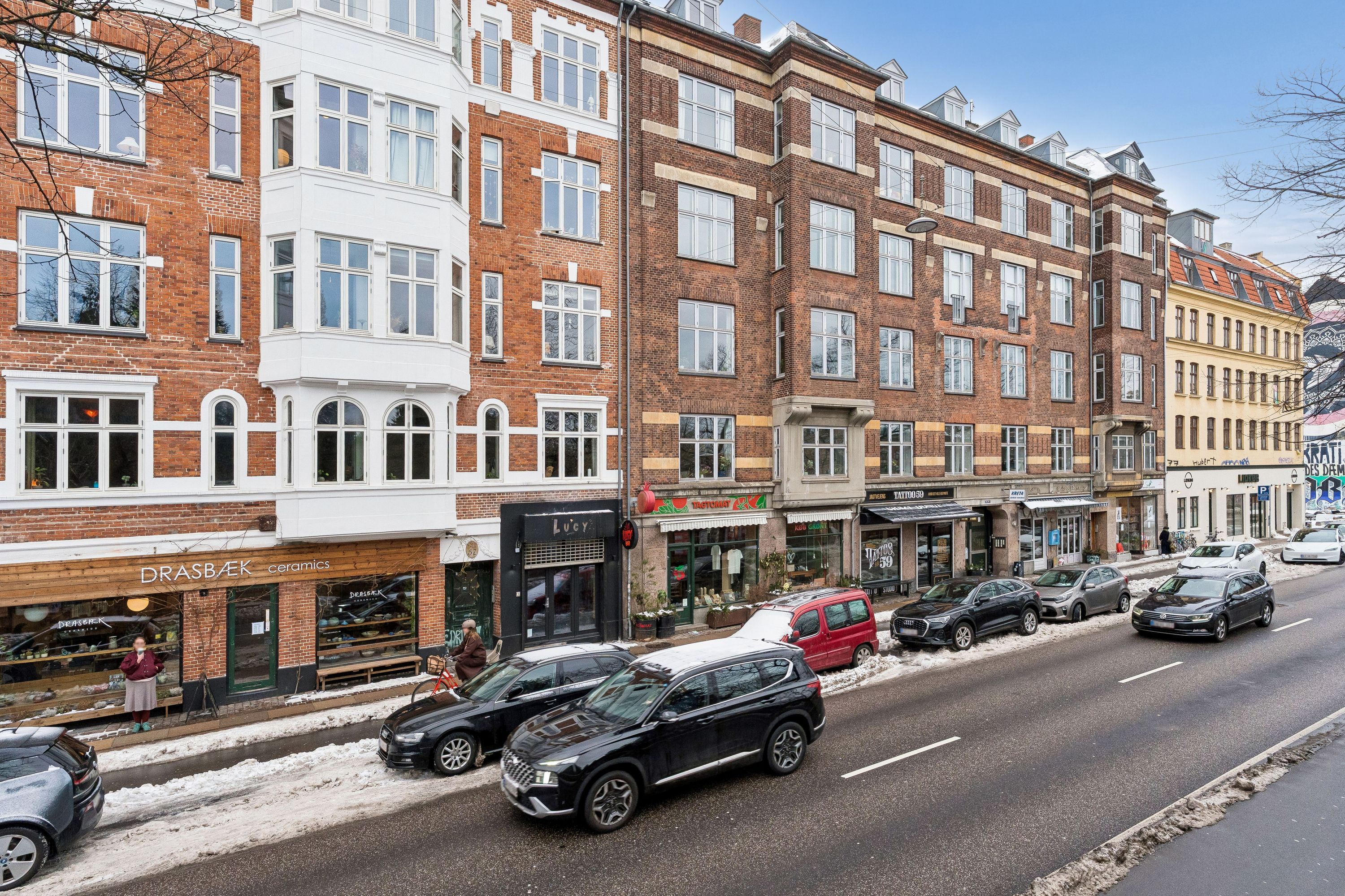 Biler parkeret langs en gågade med forretninger og folk på siden, der er sne på jorden og mennesker går forbi