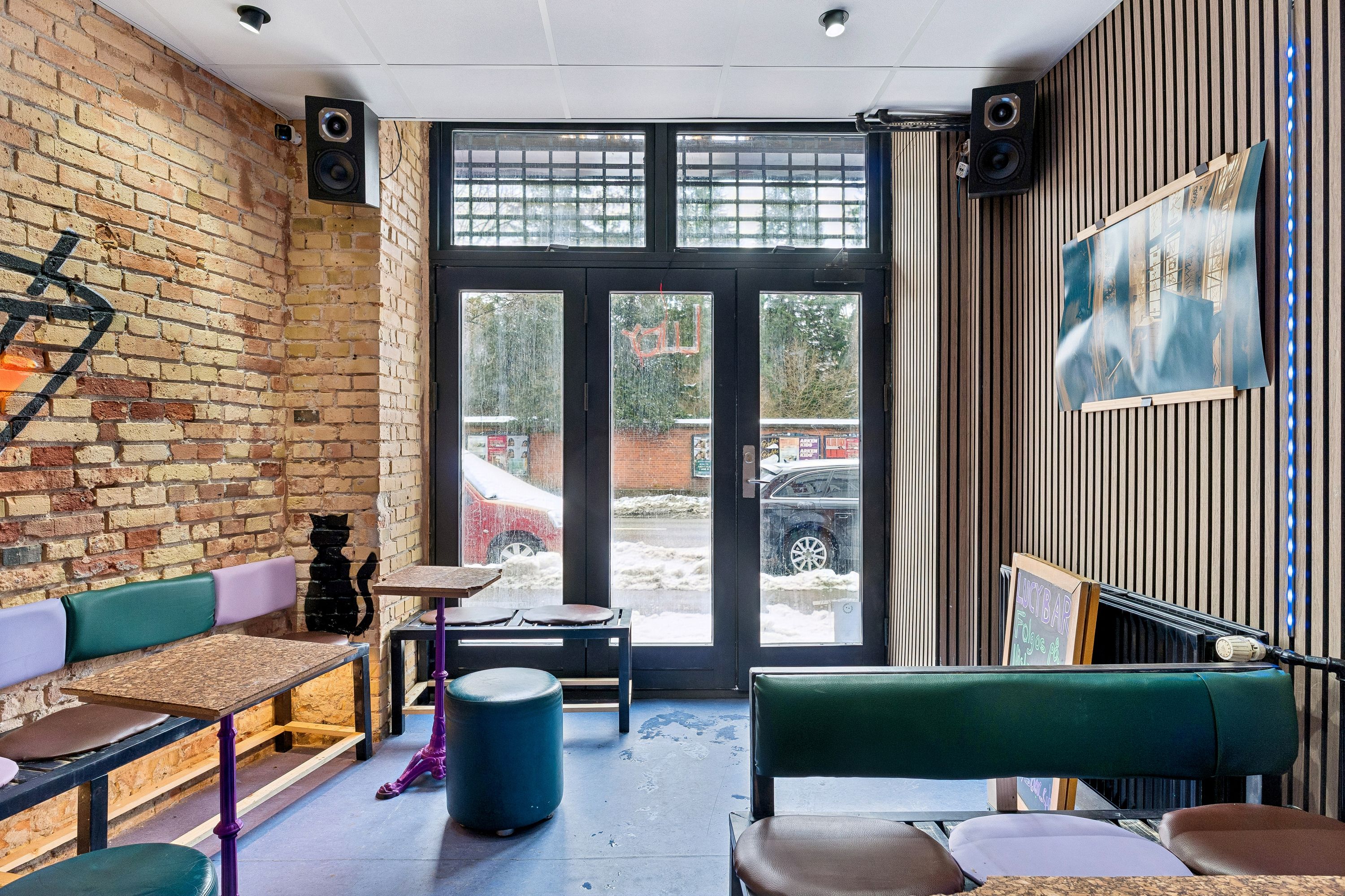 En lejet bar med grønne stole og borde samt møbler og lysanlæg i en bygning med krydsfinerbeklædte vægge og udsigt til en snefyldt vej