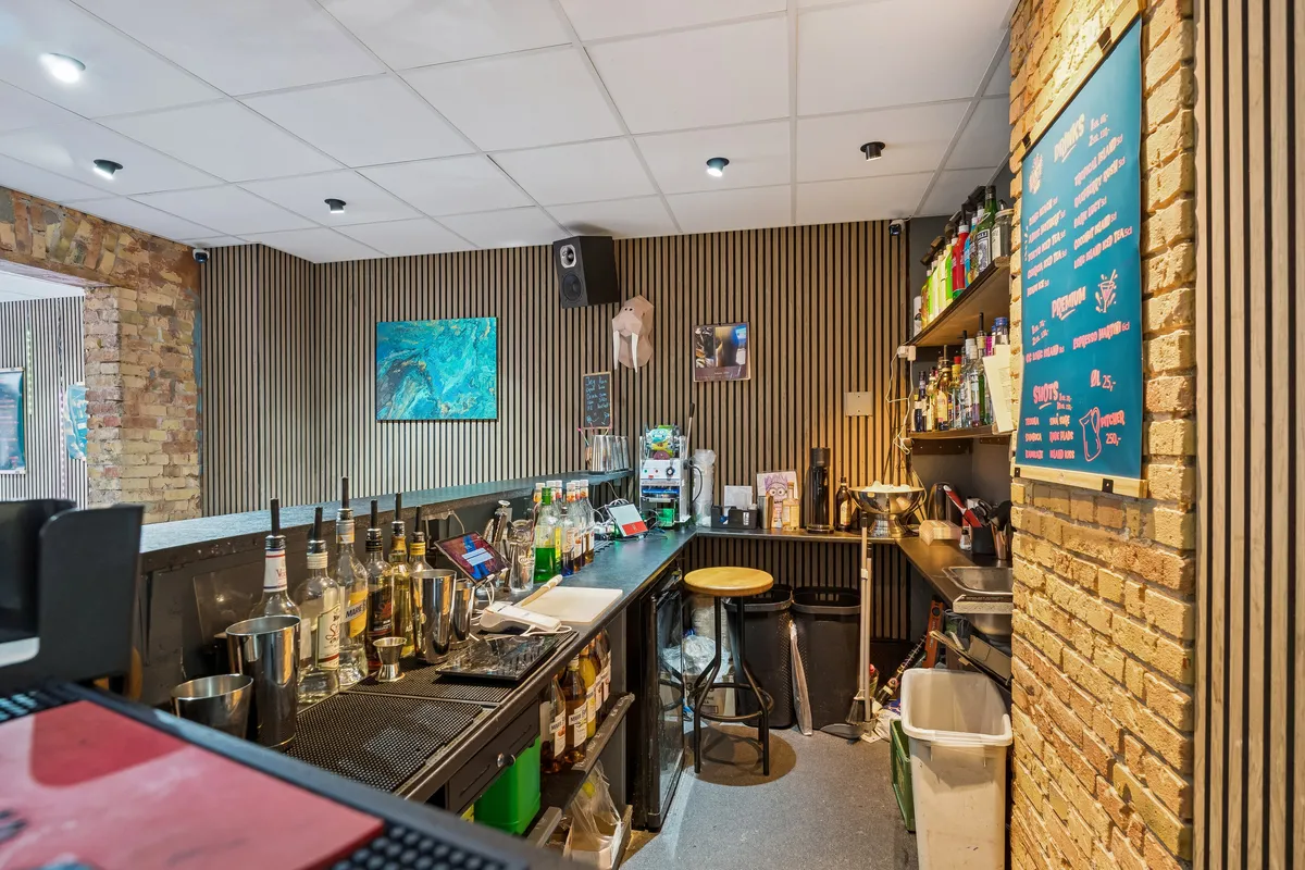 Bar med bardisk og flasker samt en menu over de drikkevarer, der serveres. Lys blytonede loft med højt til loftet og loftsmonterede lygter. Sorte søjler og gulvtæppe.