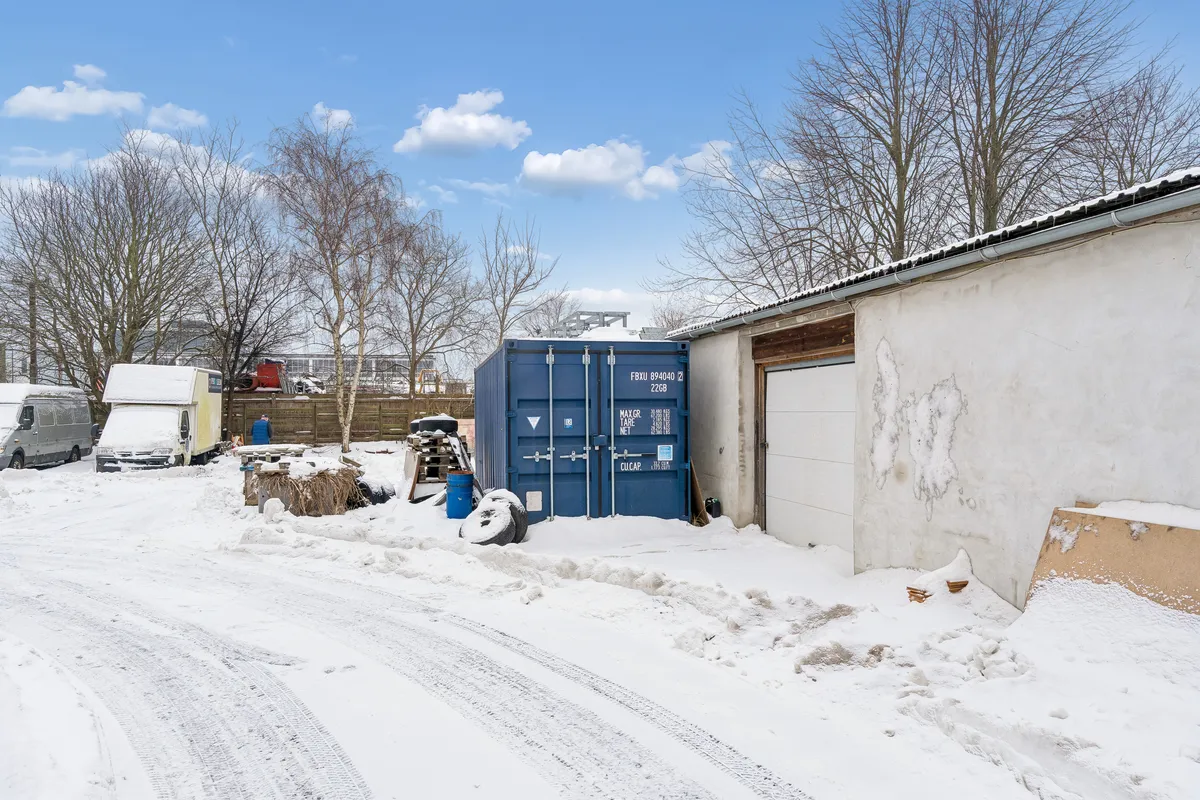 Et stort lagercontainer står på en snepakket vej med en hvid garage og en skiltet bil på den ene side og en skov på den anden side