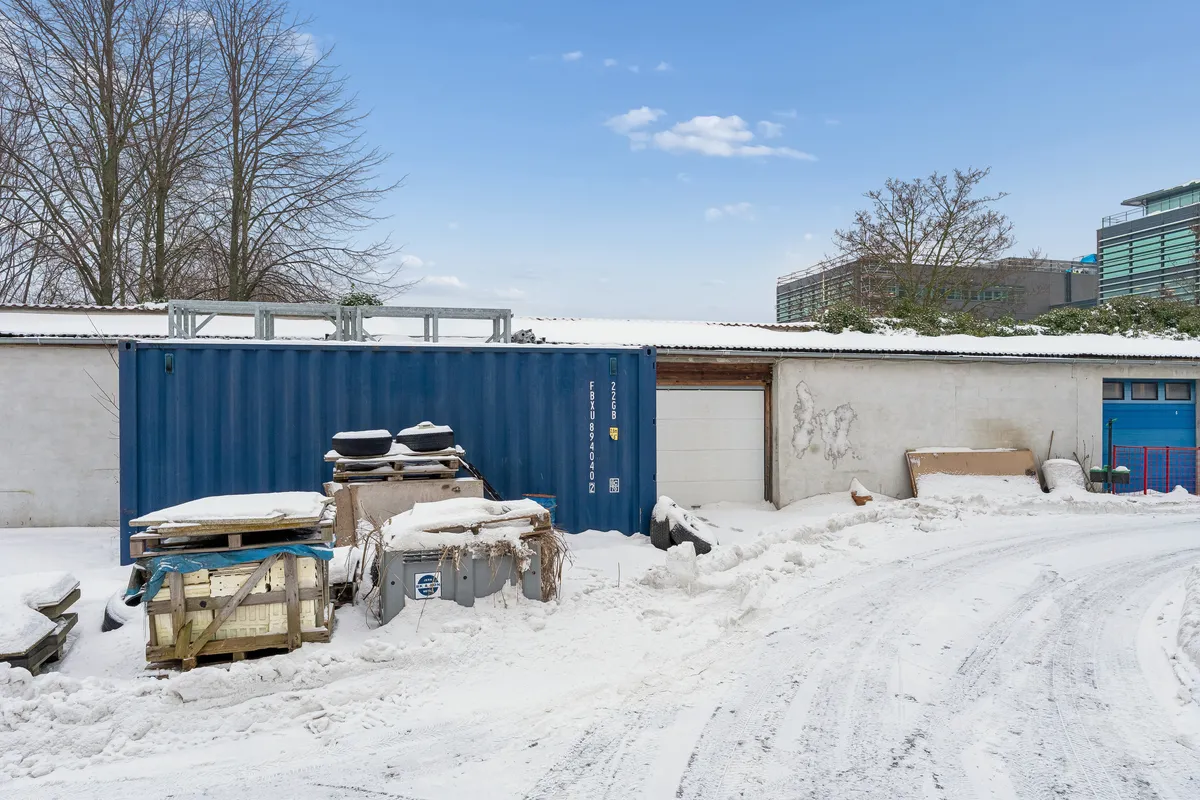 Et stort lagerhus med blå container og andet lagermateriale er omgivet af sne. Væggene er hvide og der er et port og en garagedør på huset.