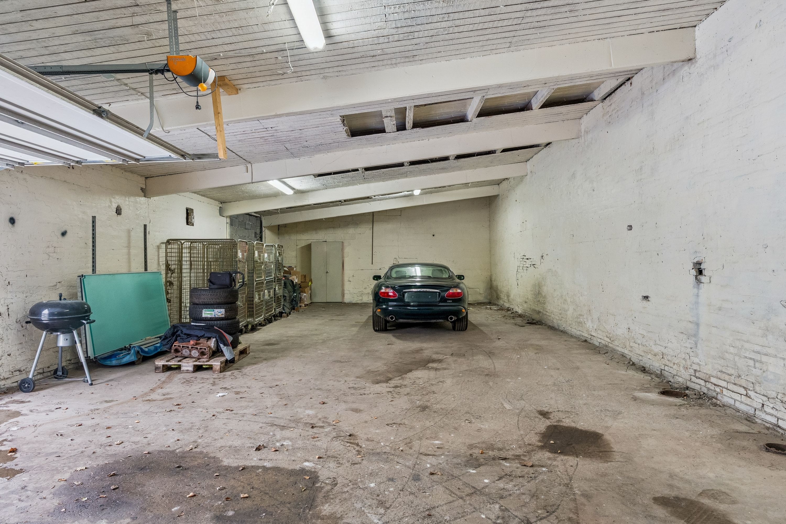 En gammel koldt lager med høj loftshøjde og mange gamle bildele og nye dæk. Der er en port og en gammel grill og en bil parkeret i enden af hallen.