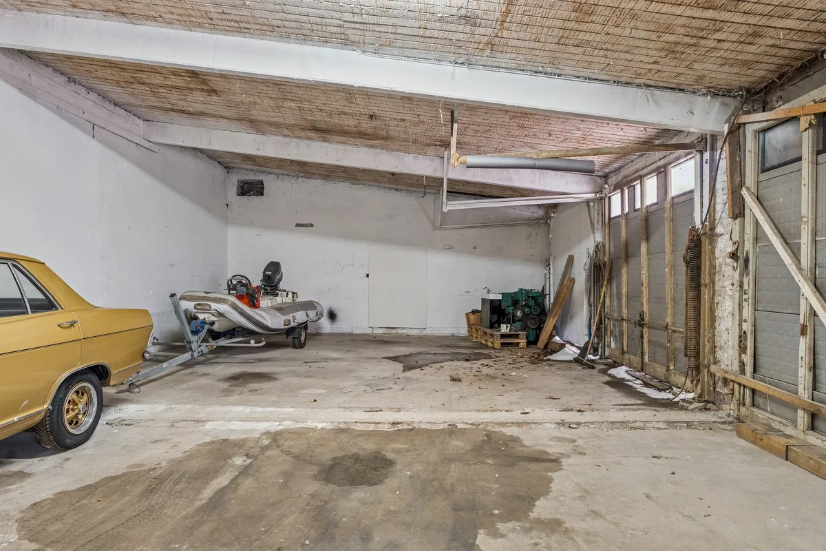 Et koldt lager med loftshøjde på ca. 3,5 meter. En gammel gullig bil og en båd på traileren. Et skab og en generator placeret på en paller.