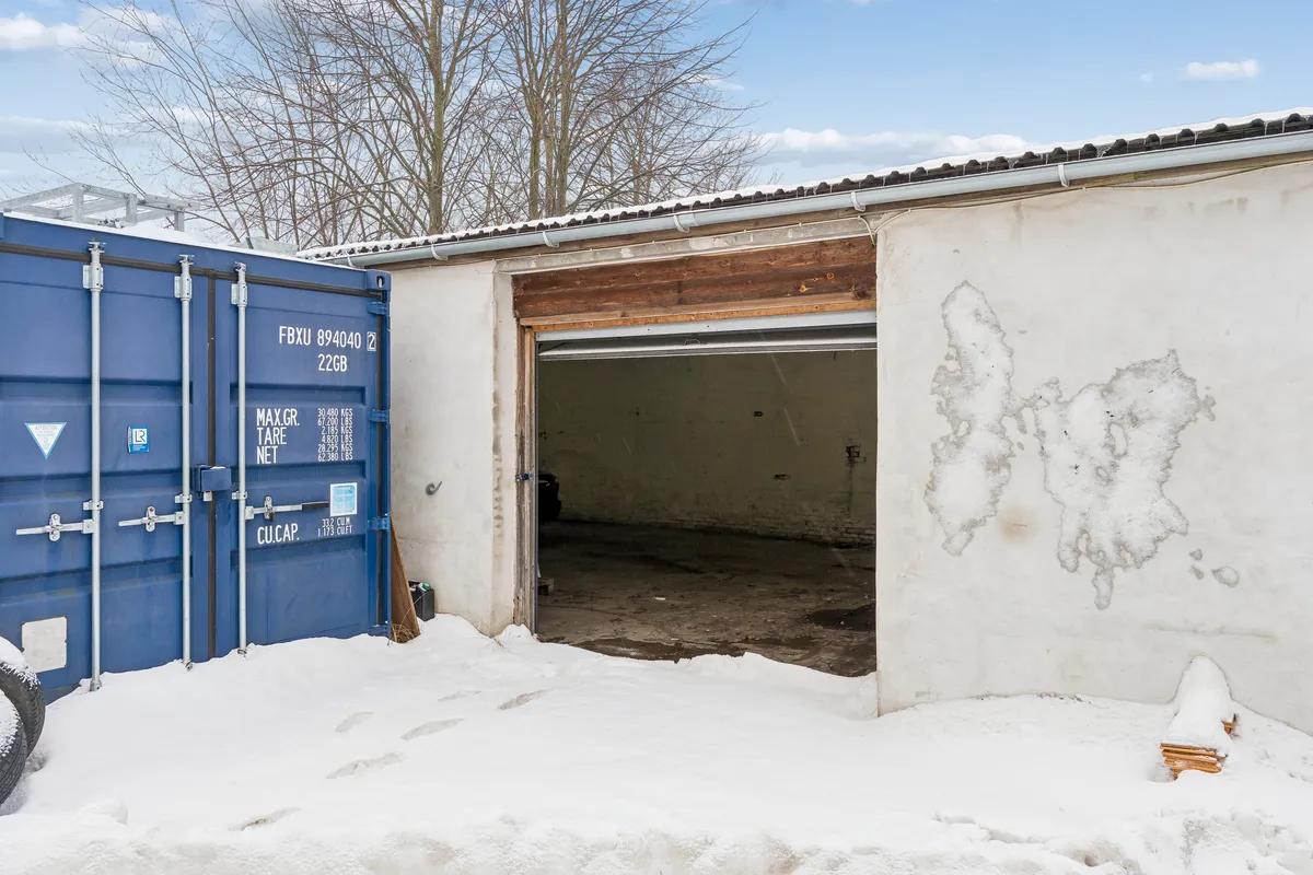 Lagerbygning med åben garagedør og sne på jorden, en stor blå container står ved siden af, og der er spor af sne på væggen
