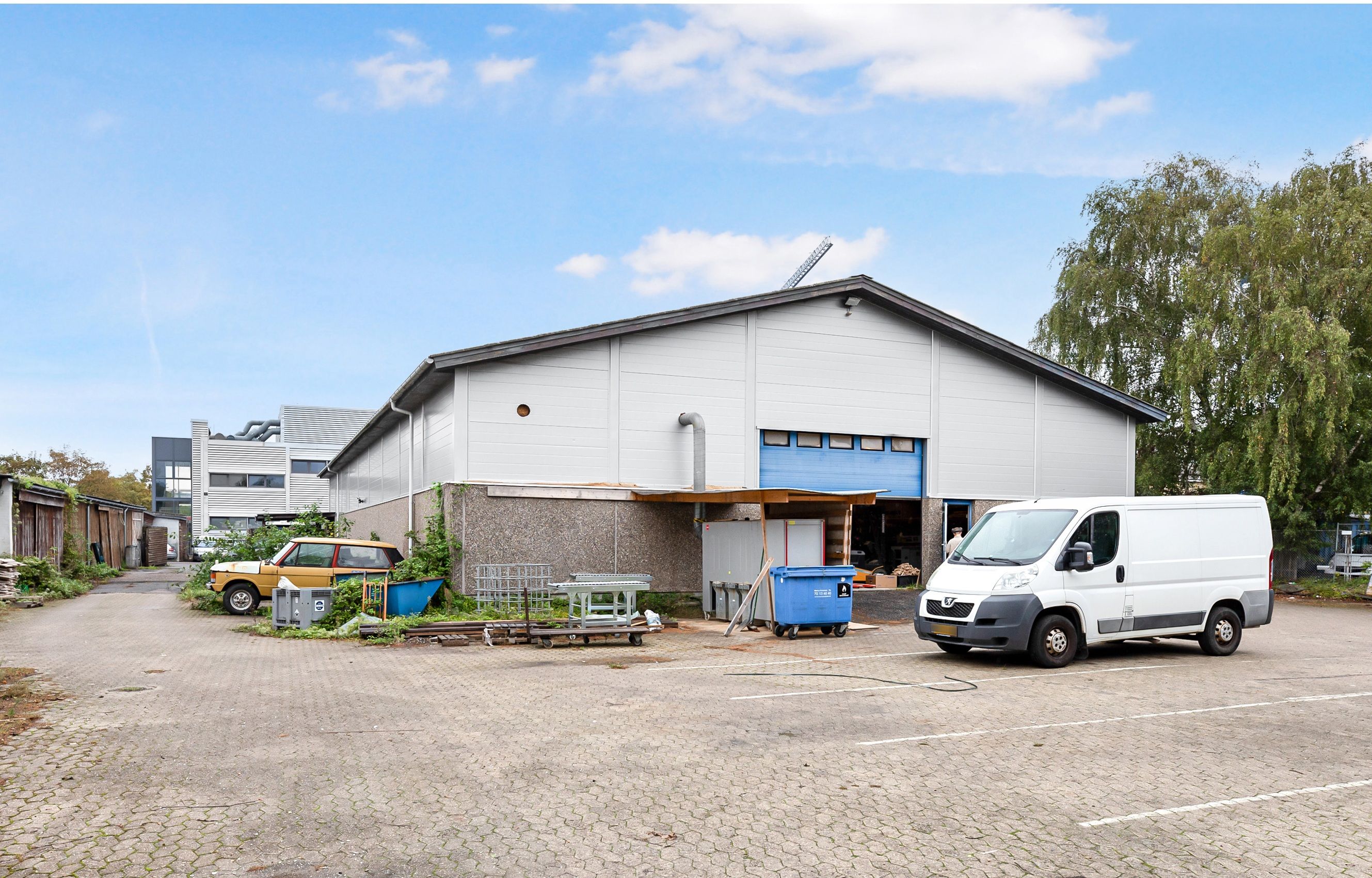 Et stort lagerhus med en blå port og en hvid kassevogn parkeret foran og en gul bil parkeret på siden af bygningen