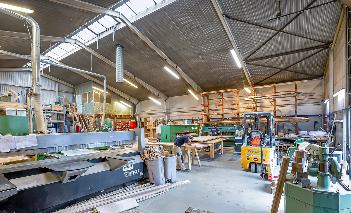 Lagerbygning til leje med flere maskiner og materialer, flere arbejdsborde, højt loft med stålbælte og lysarme, og flere mænd står og arbejder