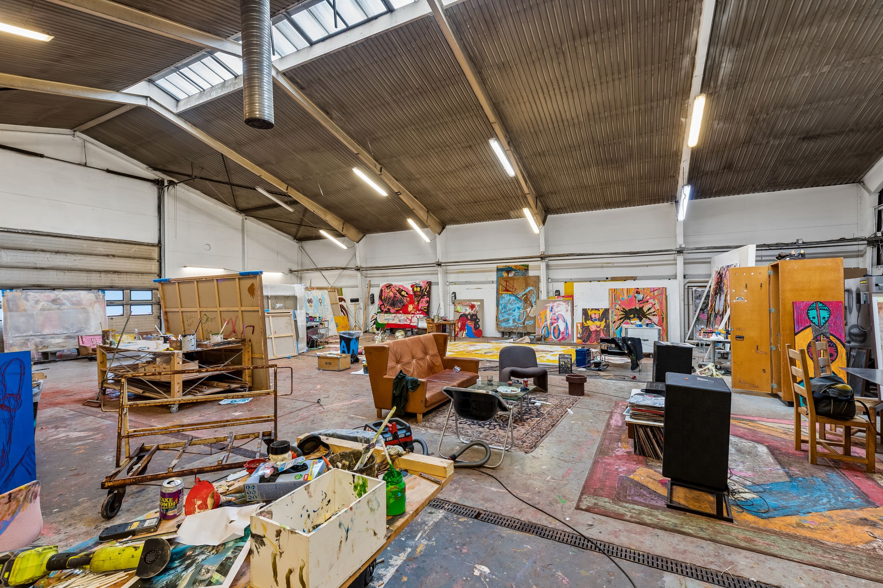 Malerværksted med flere store malerier og værktøj i en stor lejehal med højt loft og store vinduer