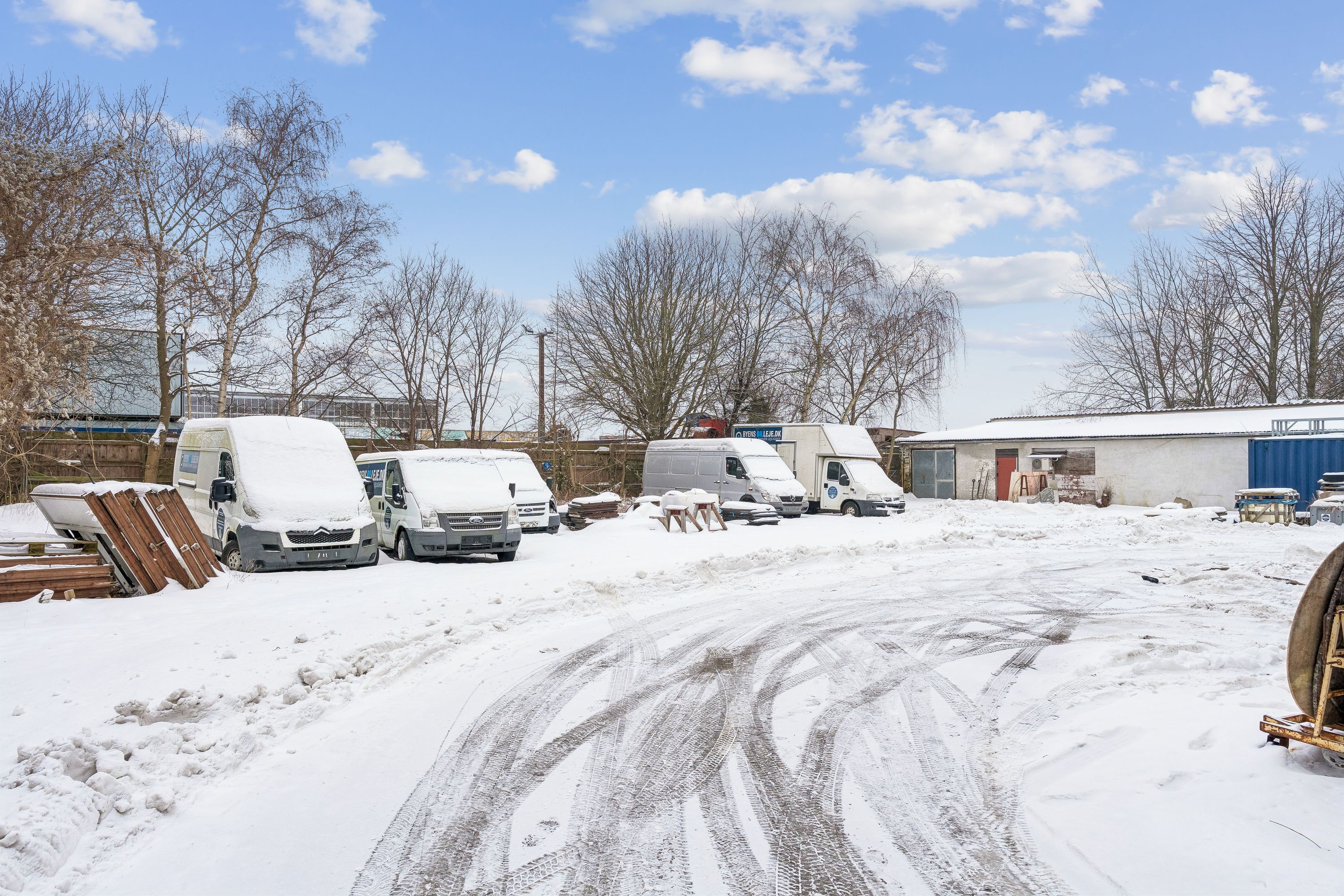 En sne-dækket vej med flere parkerede erhvervsvogne og bygninger, og sne på træer og tag
