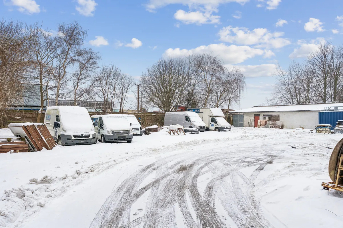 En sne-dækket vej med flere parkerede erhvervsvogne og bygninger, og sne på træer og tag