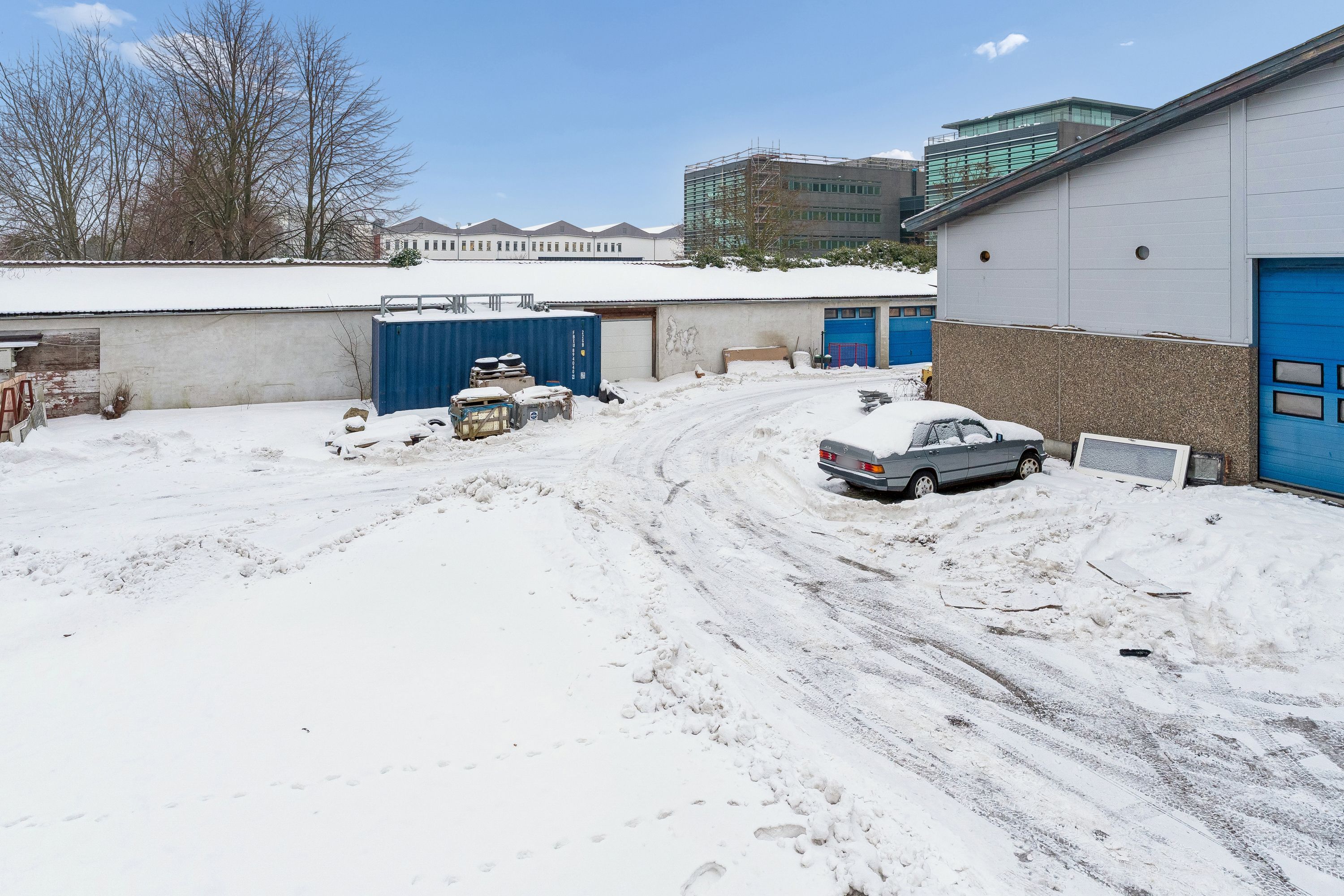 En gammel bil parkeret i sne, gamle og nye bygninger med snedækkede tag, og mange træer i baggrunden.