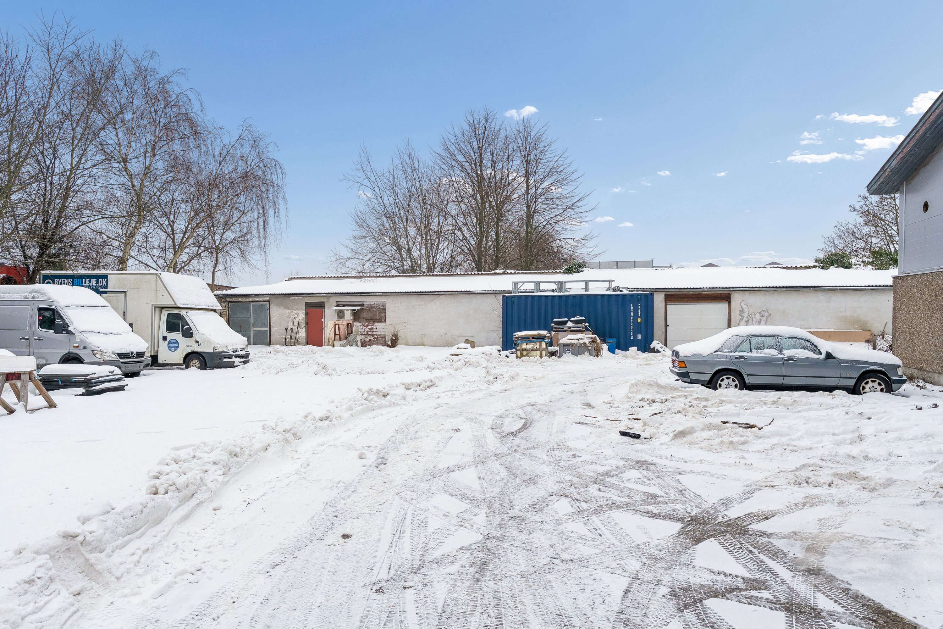Hvidt sne dækker vejen mellem biler og bygninger med skilte, og træer uden løv.