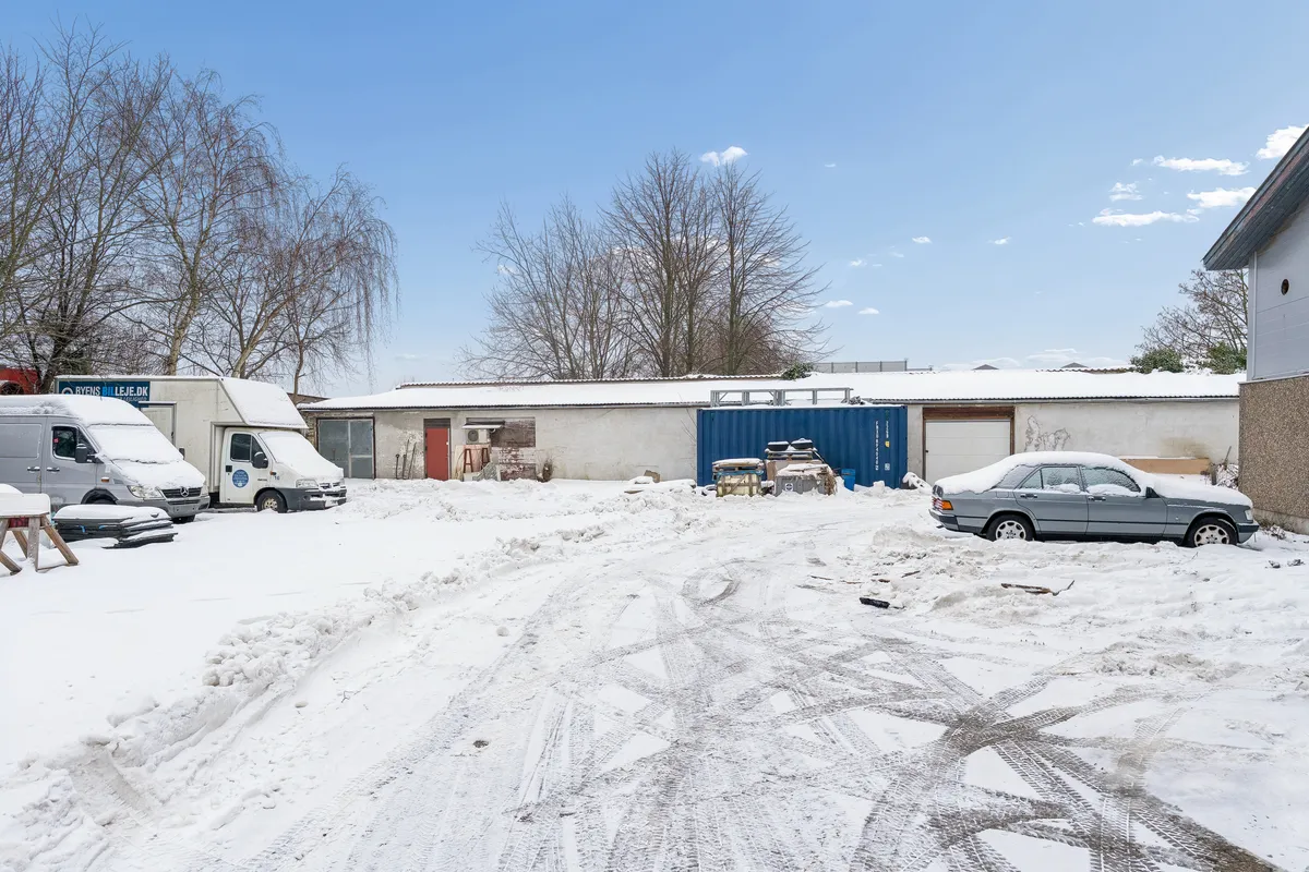 Hvidt sne dækker vejen mellem biler og bygninger med skilte, og træer uden løv.