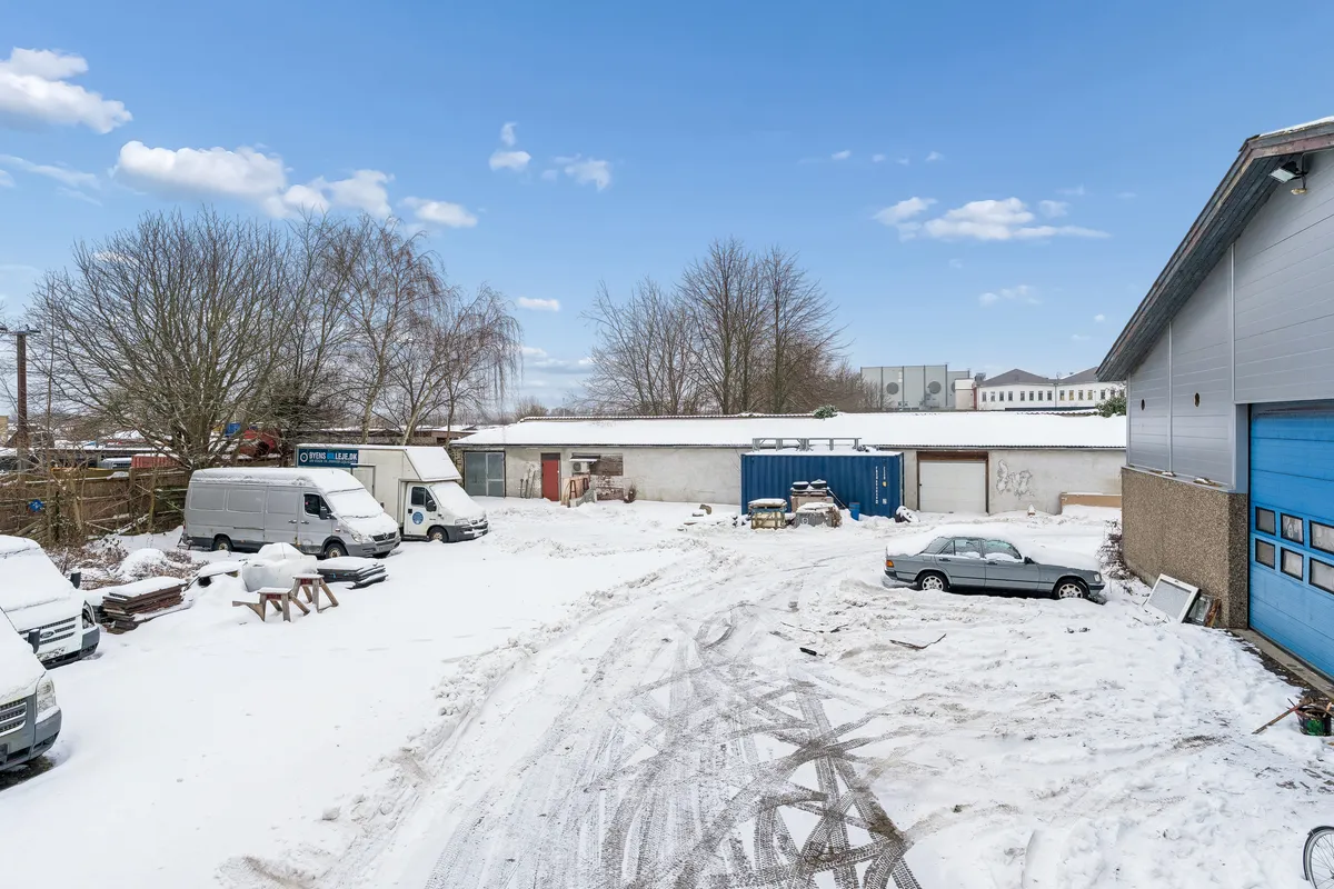 Industrielt område med flere lastvogne og biler i sne, garager og bygninger med blå døre, træer og bygninger i baggrunden