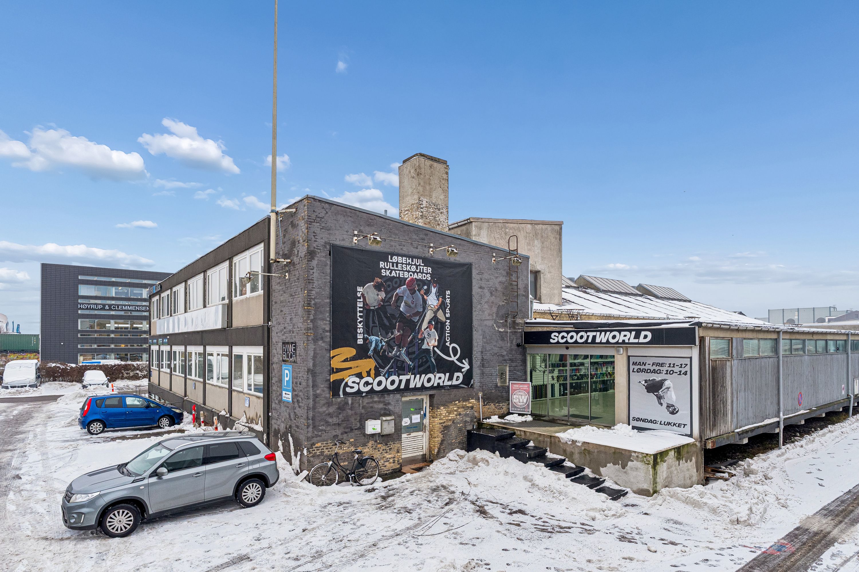 Lagerbygning med Scootworld skilt og skilte for åbningstider, parkeringsplads med to biler, og en cykel udenfor bygningen, alt sammen dækket af sne.