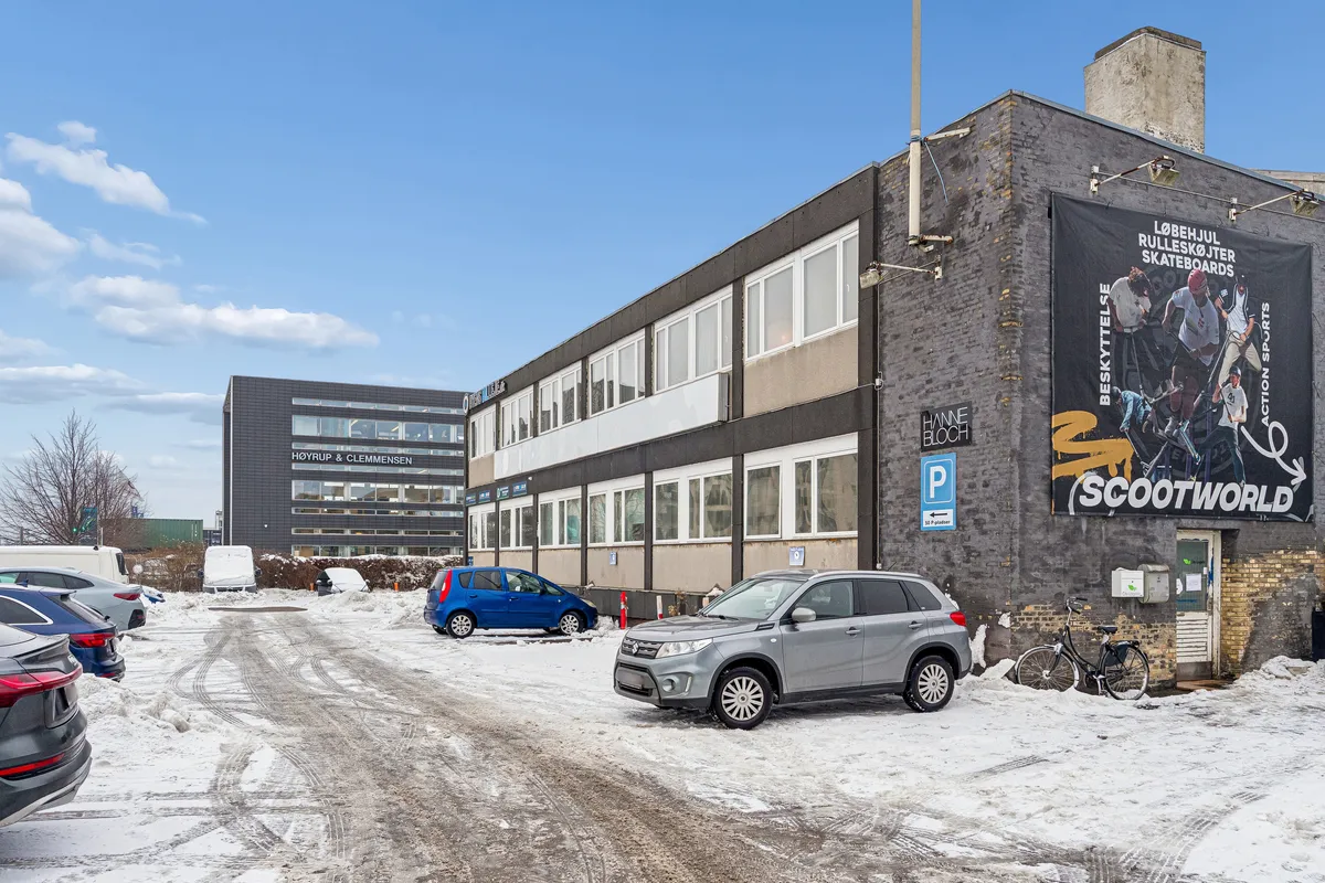 To biler parkeret foran bygning med skilt og skilte med tekst og logoer i sne, vinduet og højt opbygget facade, sne og en cykel står udenfor døren