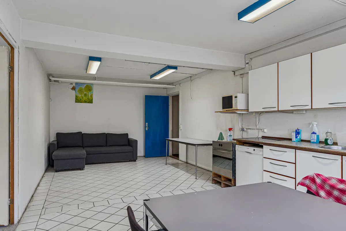 En funktionel og lys kælderlejlighed med højt loft og hvide vægge, en sofa, køkken med ovn og et stort skrivebord med stole og en træpalle