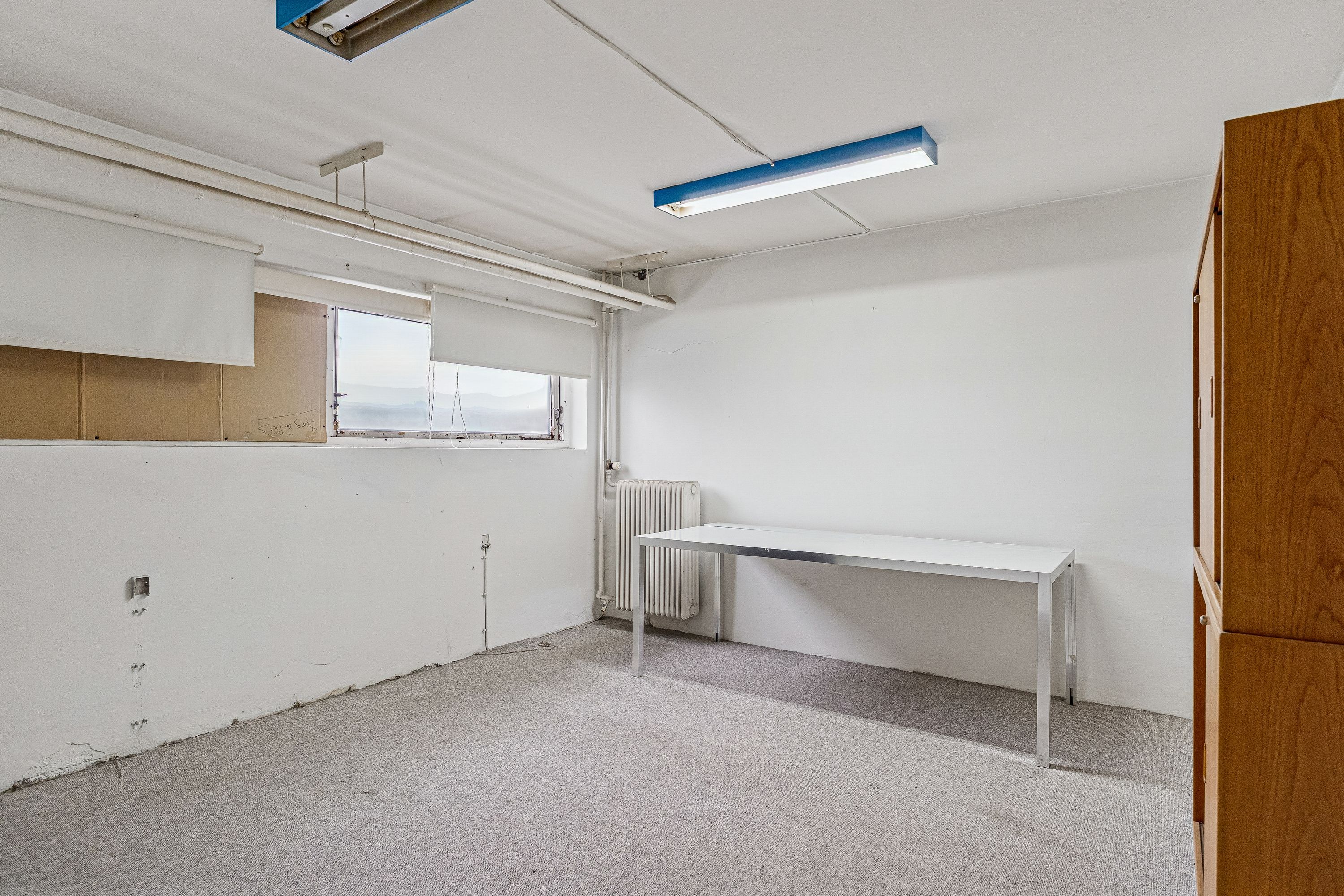 En tom, lysegrå lagerrum med hvide vægge, en hvid bord og en hvid radiator samt to store vinduer med hvide gardiner i baggrunden