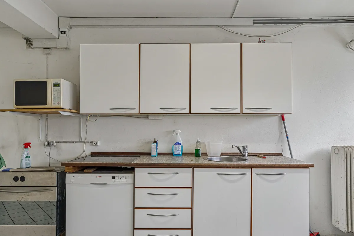 Kælderlejlighed med køkken med hvide skabe og lysegrå loft. Ovn og mikrobølgeovn er installeret, og der er lysegrå, rustfri strygejern og en hvid radiator.