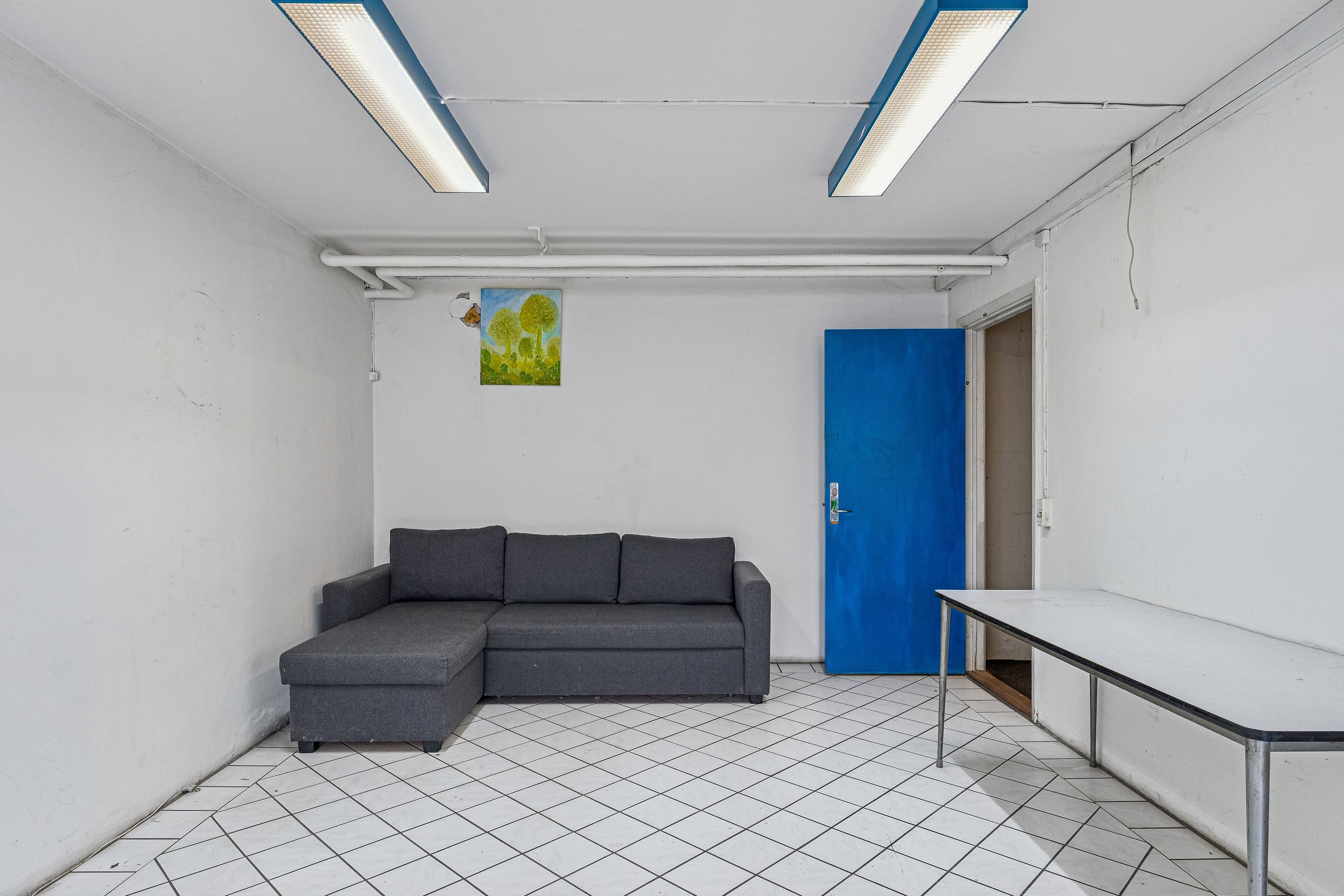 Lager med højloft og lyse loftslamper, funktionelt sovesofa og hvidt køkkenbord, hvor gulvet er klædt med hvide kvadratiske fliser og en lyseblå dør