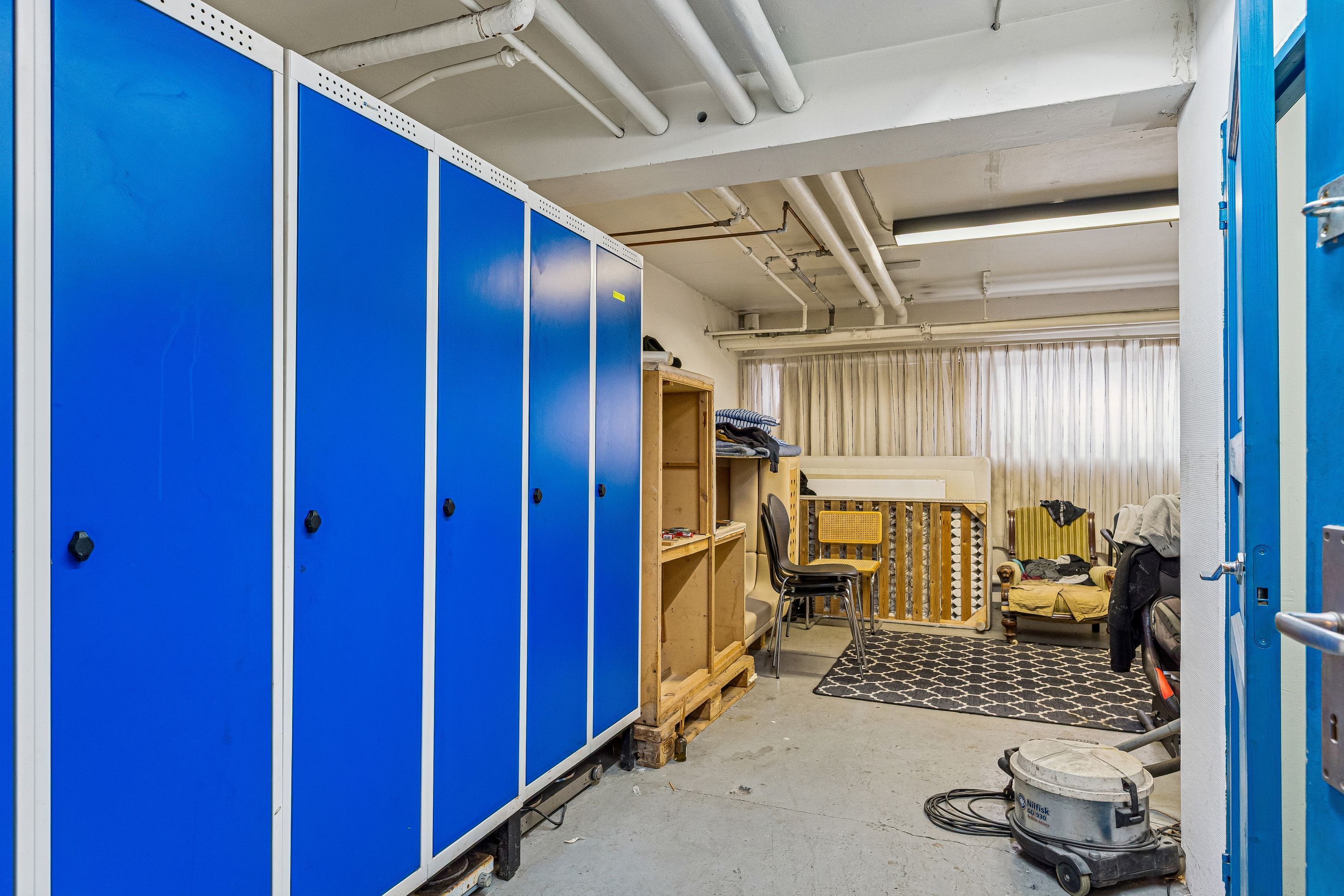 Lager med blå metalskabe og højloft, lyse rullegardiner og gulvtæppe, tøj på bænk og stol, støvsuger og træpalle