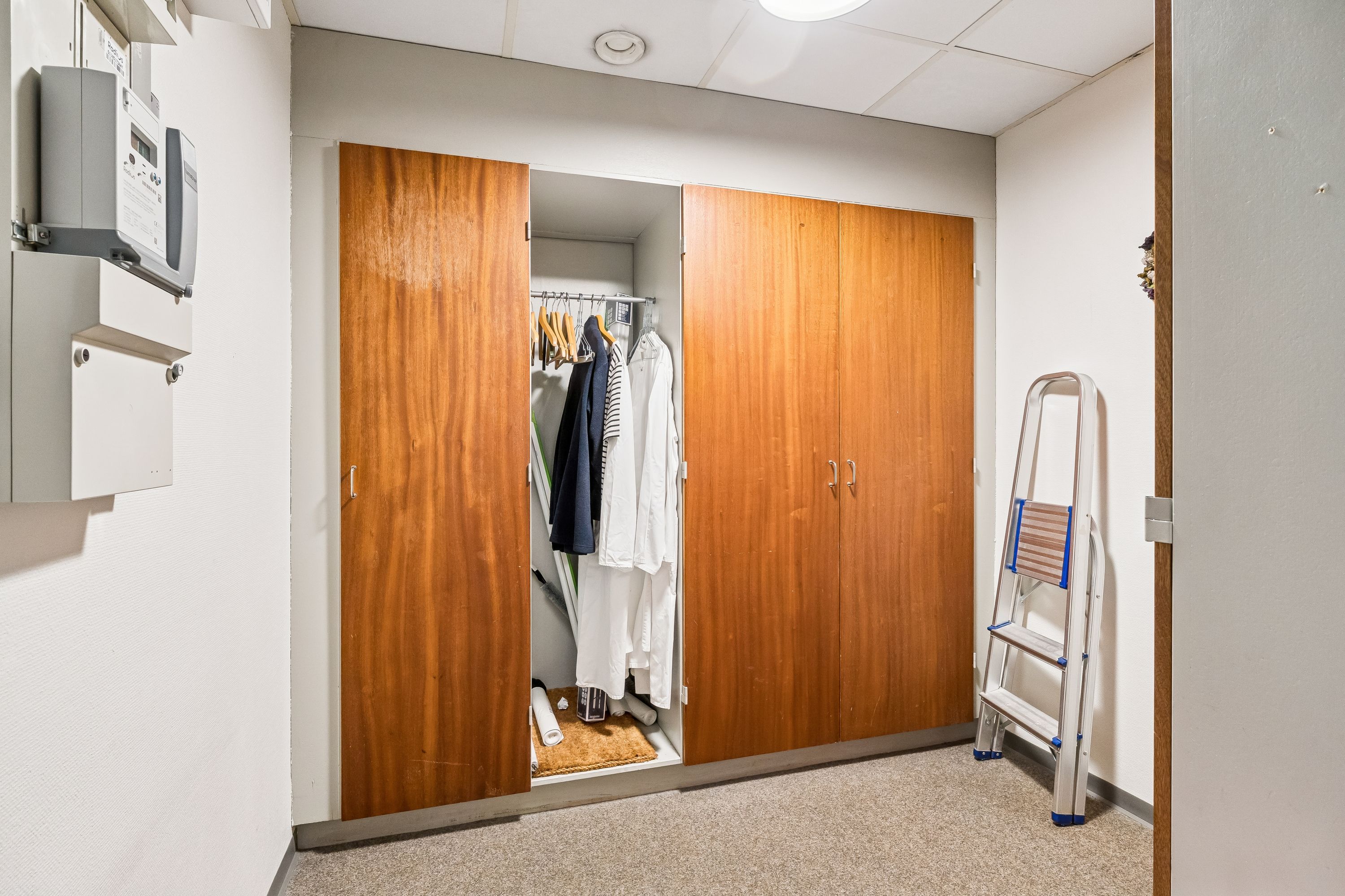 En åben garderobe med to store trædøre og indvendigt hyldesystem, samt en stående stige mod højre. Inde i garderoben er der hængegrej og tøj.