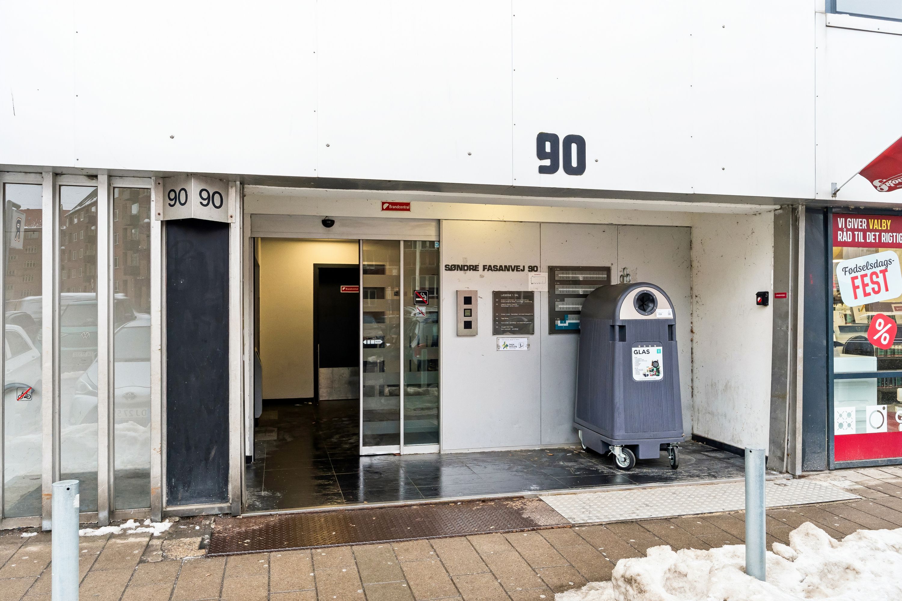 En bygning med indgangsdøren åben og nummeret 90 på facaden. En skilt med teksten Søndre Fasanvej 90. En høj, sort affaldscontainer med hjul og en reklameskilt med teksten Fest