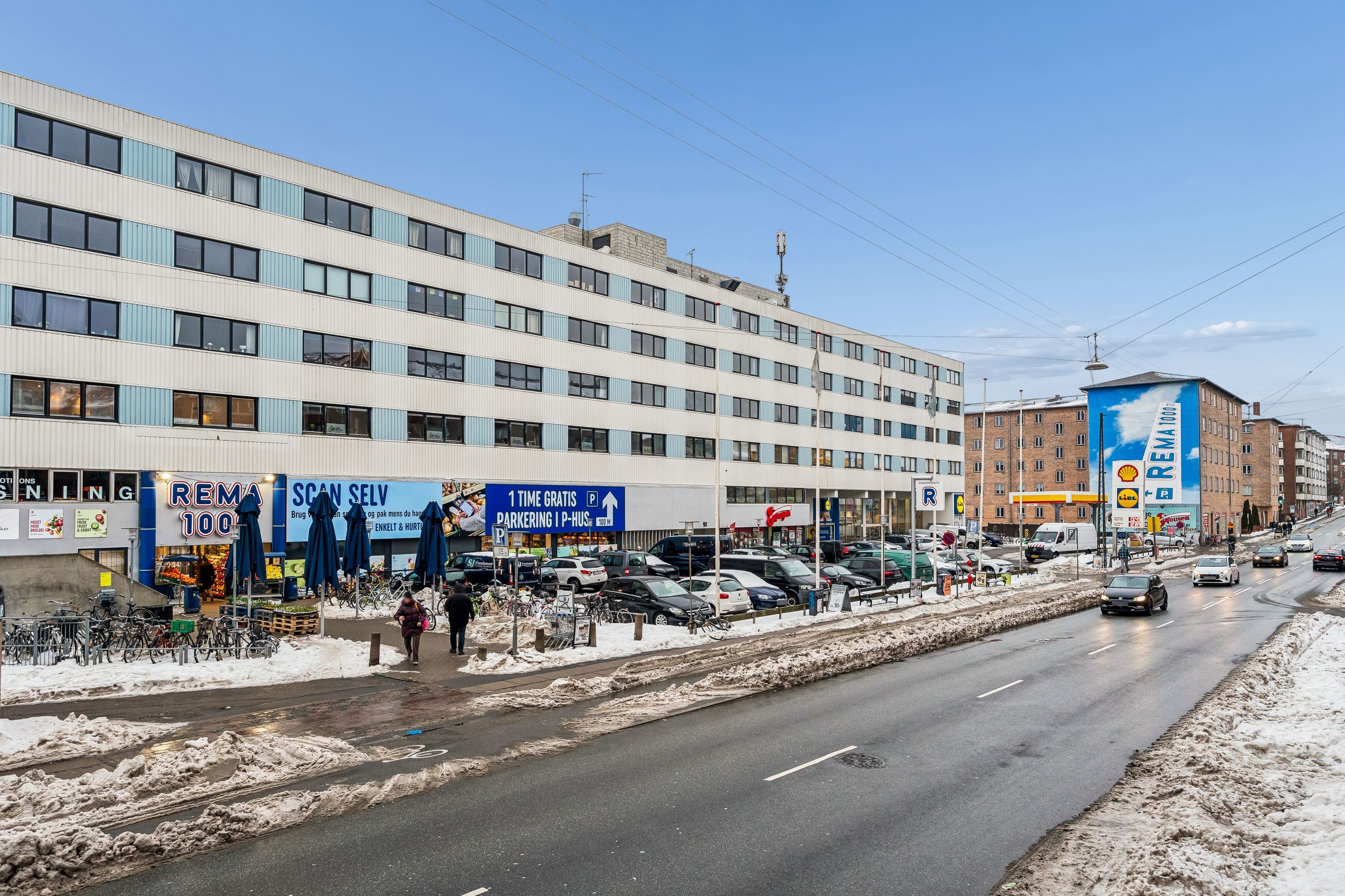 Kontorbyggeri med mange butikker og parkeringspladser samt gågade og biler i forgrunden, sne på jorden og vejen og biler kører på vejen
