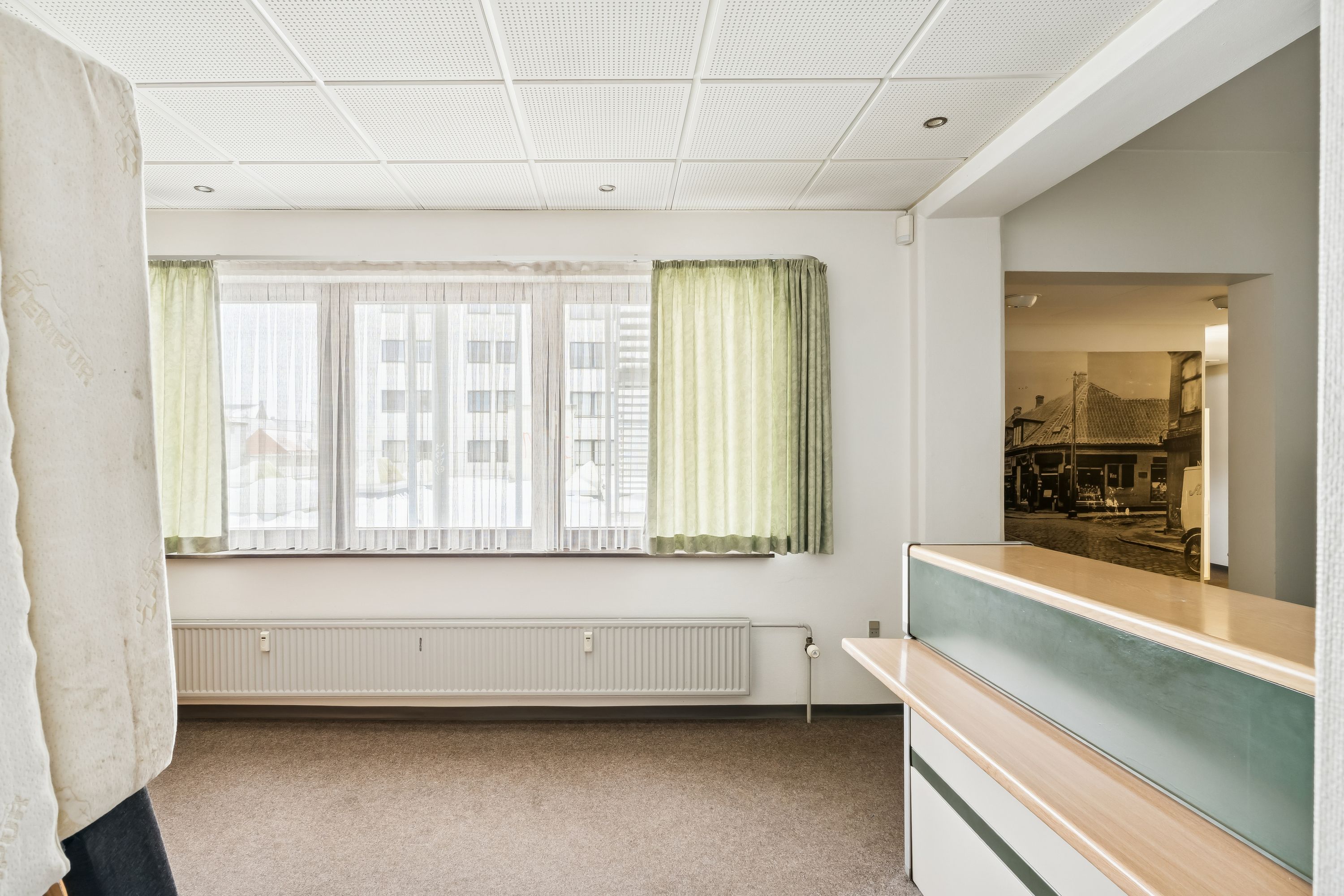 Kontor med slanke linjer, moderne møbler, hvide vægge og lyse gardiner, et radiator, et lysekron og et skrivebord med en lysegrå kant.