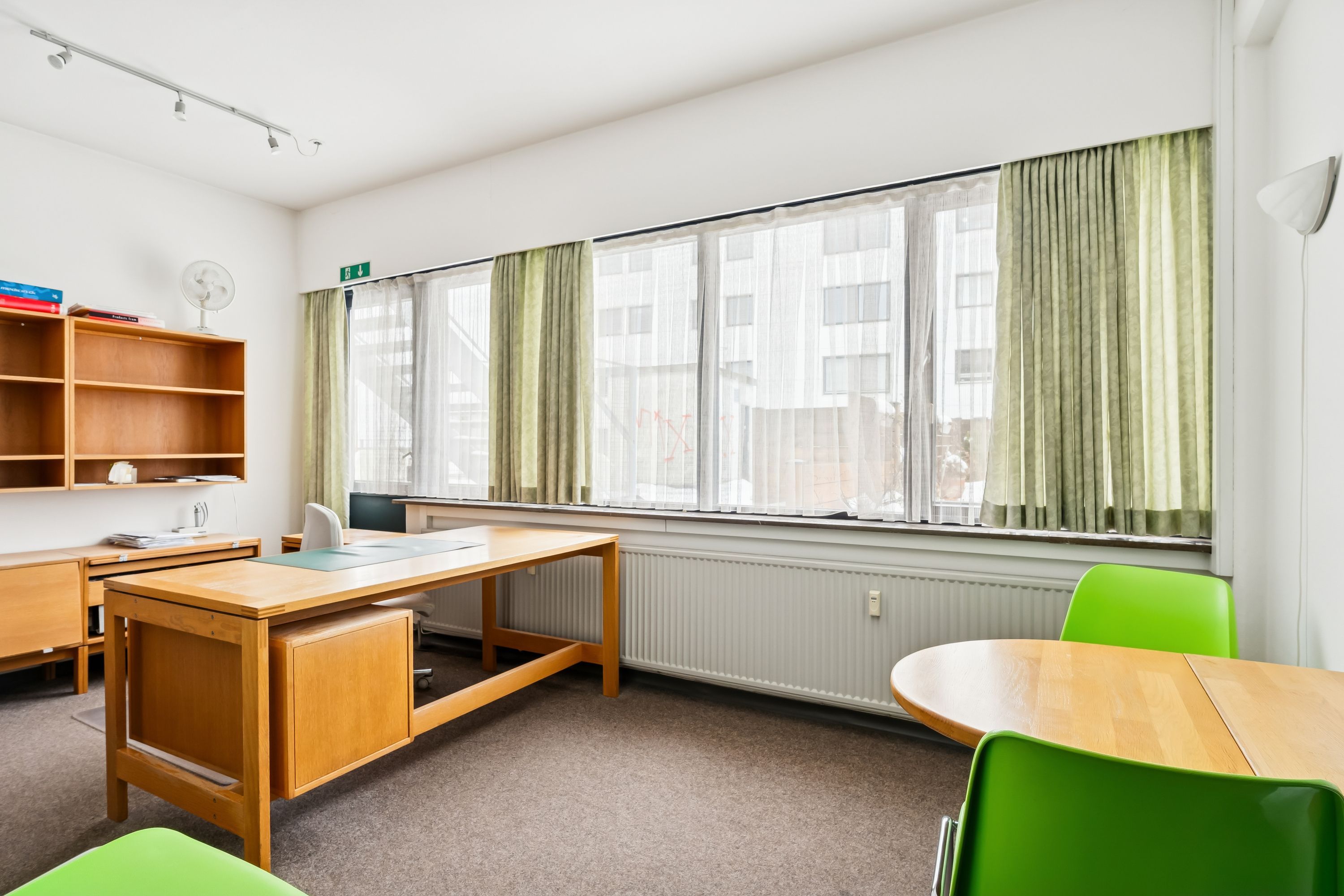 Kontorrum med lyse møbler, en stor skrivebord, grønne stole, hvide gardiner og en radiator nedenunder vinduet
