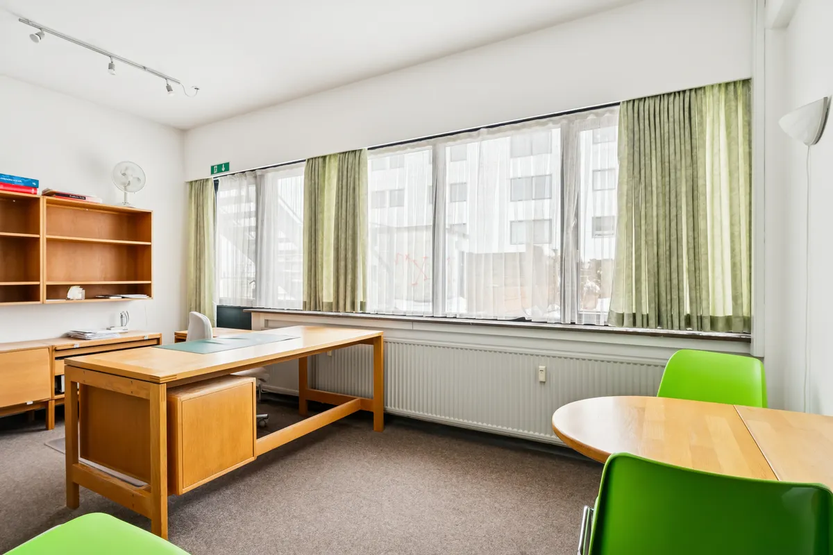 Kontorrum med lyse møbler, en stor skrivebord, grønne stole, hvide gardiner og en radiator nedenunder vinduet