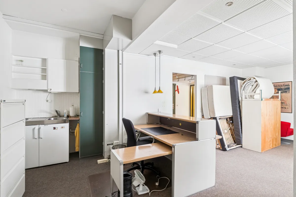 Kontor med lyse farver og moderne møbler, højloft og lyst køkken med hvide kakel og grønne kabinetter samt loftslys, der skaber en åben og indbydende atmosfære