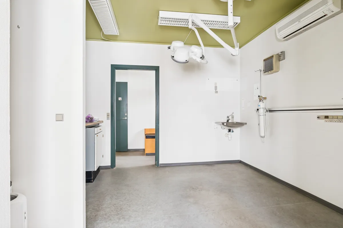 Et renseri med åbent område, stol og håndvask. En grøn dør og et hvidt loft med luftaffugter. En gul stol og en skraldespand med en hvid væg.