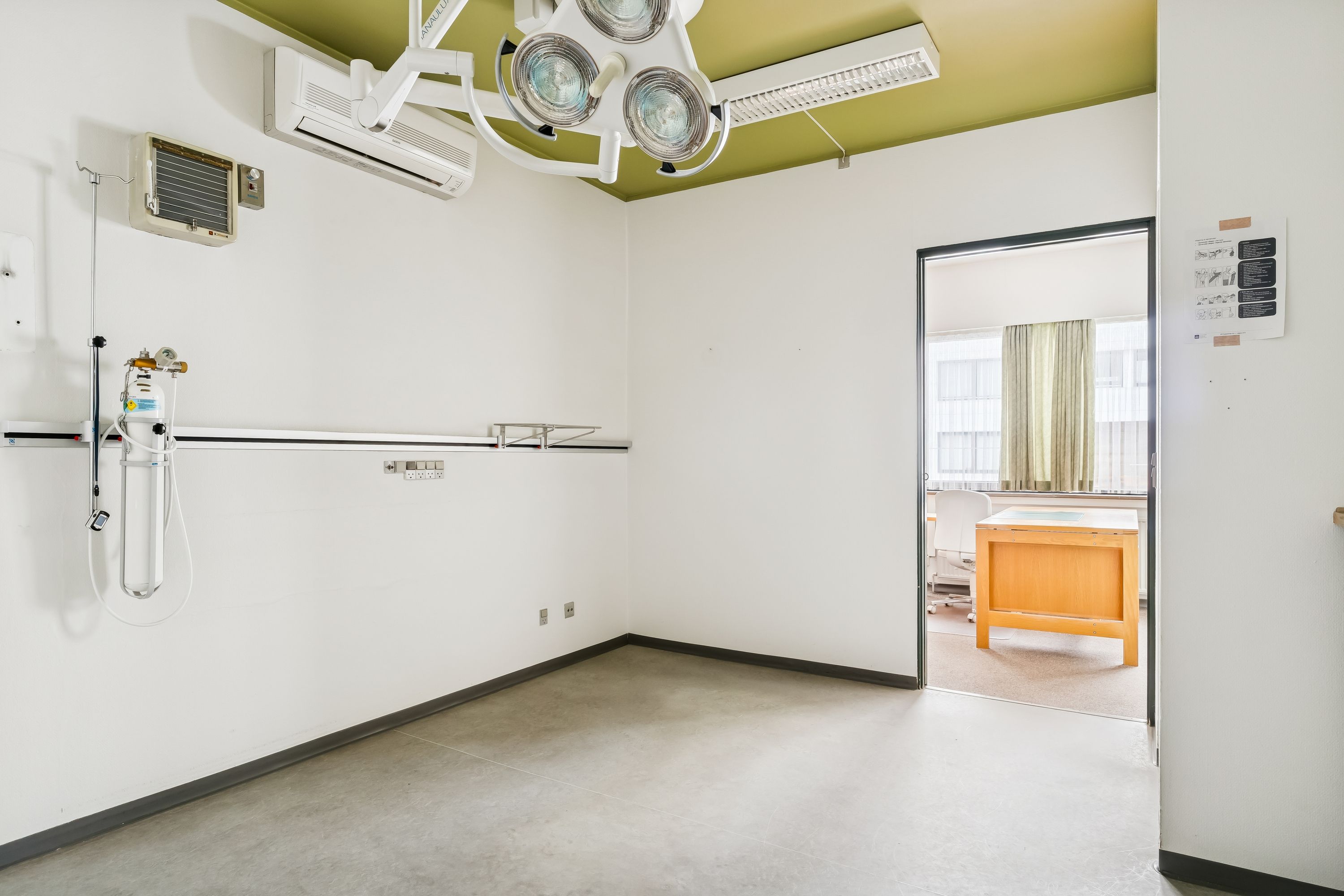 En tom, hvidvægget kontorlokale med en lysekron, en sengebord, en hængeovnblæser og et lægebord med en hvid stol og en gul gardin i baggrunden