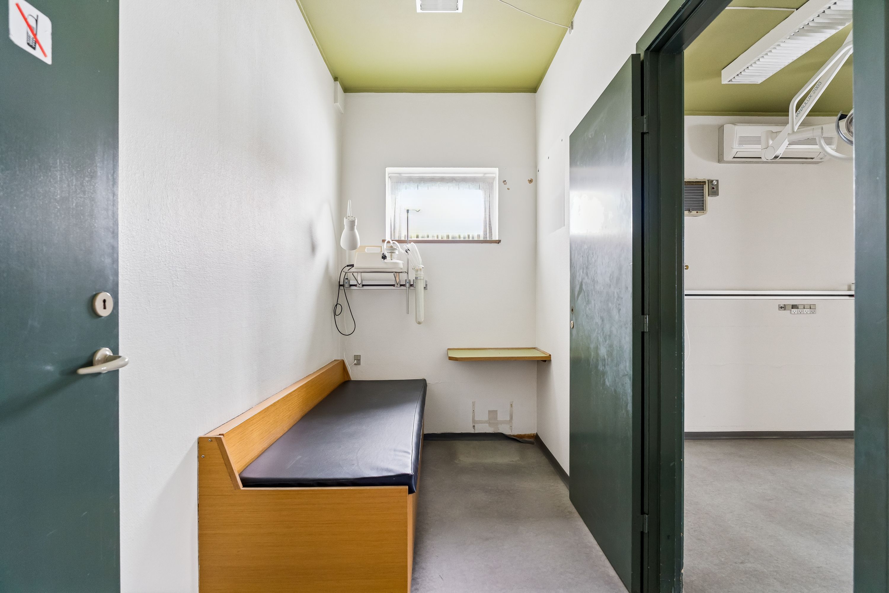 Kontorrum med dobbeltdør, hvidt vægpanel og lysegult loft, indretning inkluderer en lænestol og en lille skrivebord med en lampeskærm, og en smal vinduesåbning med udsigt til gaden