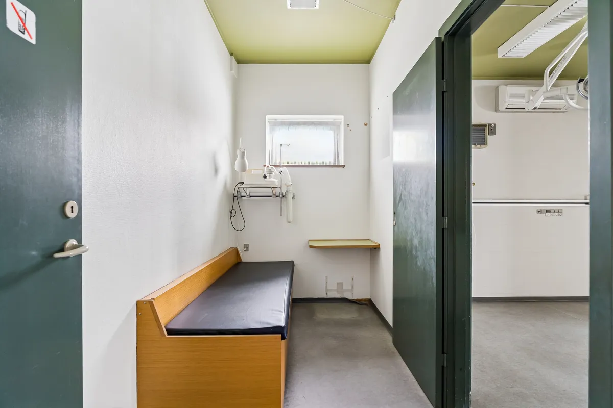 Kontorrum med dobbeltdør, hvidt vægpanel og lysegult loft, indretning inkluderer en lænestol og en lille skrivebord med en lampeskærm, og en smal vinduesåbning med udsigt til gaden