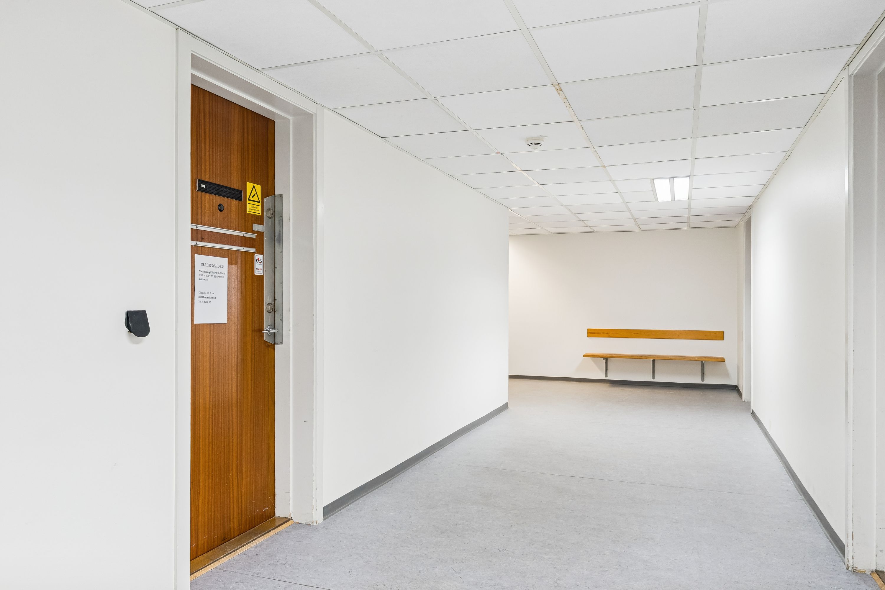 En tom korridor med hvide vægge og lofter og en brun dør med adgangskode. Der er et gult skilt og et hvidt papir med tekst på døren. En smal bænk står mod væggen